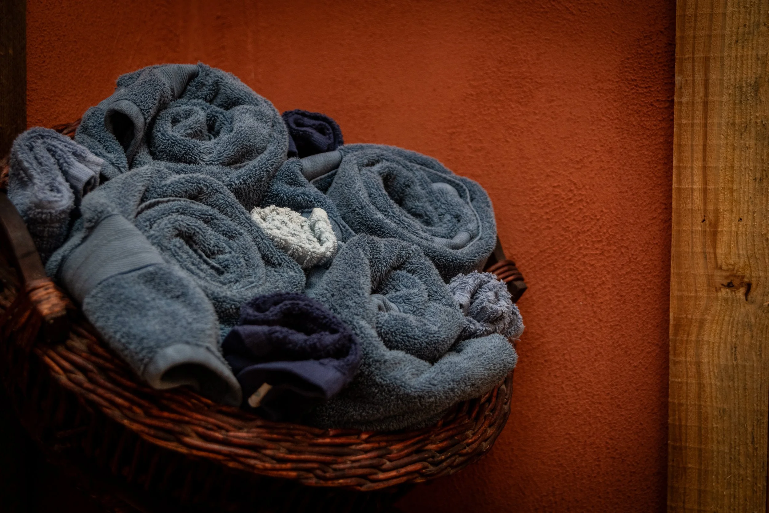 A basket filled with rolled and folded blue towels placed against a textured dark orange wall with a wooden post visible on the right side.