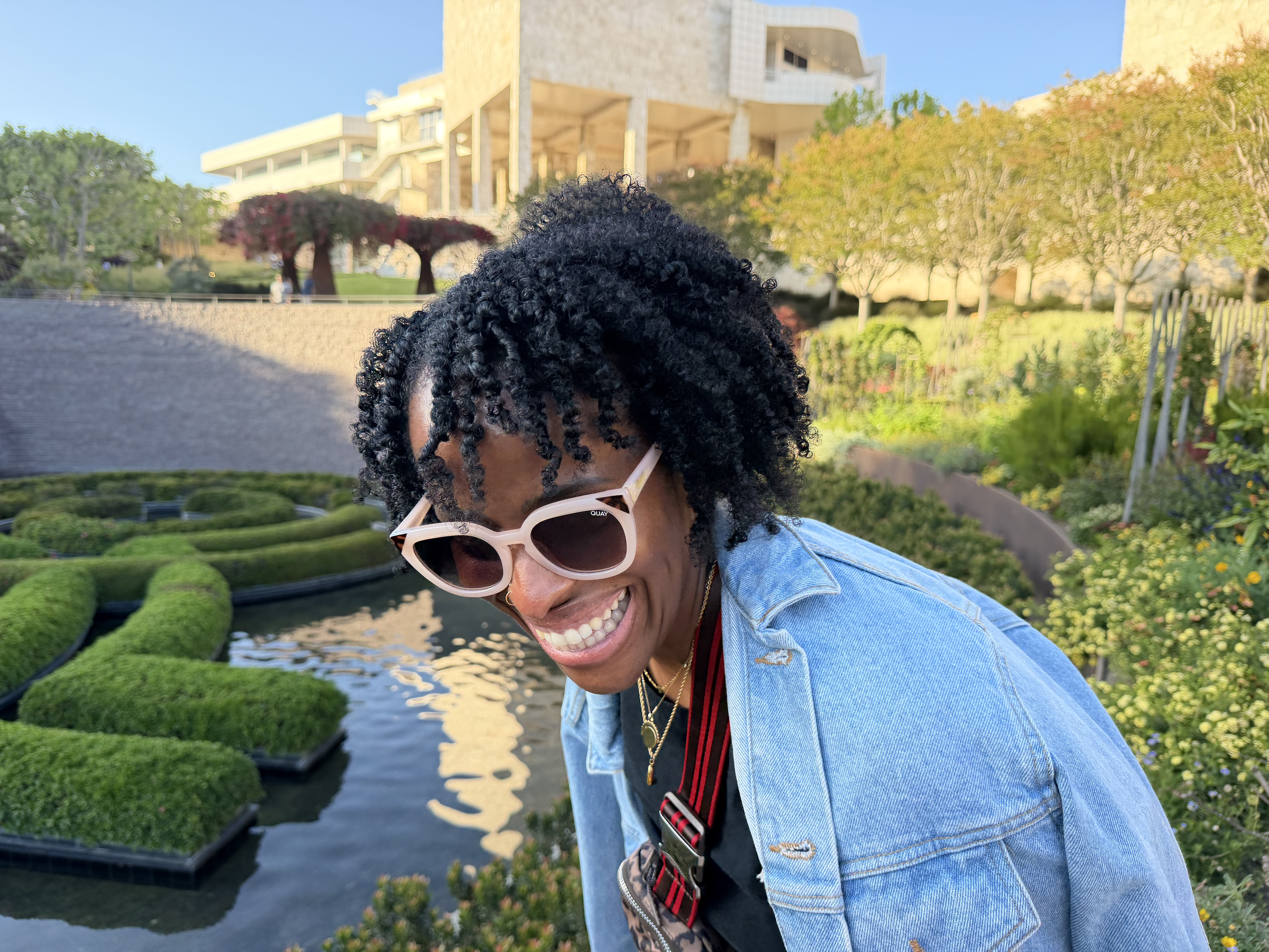 Smiling woman with curly hair wearing sunglasses and denim jacket in garden with trimmed bushes and modern building in background.