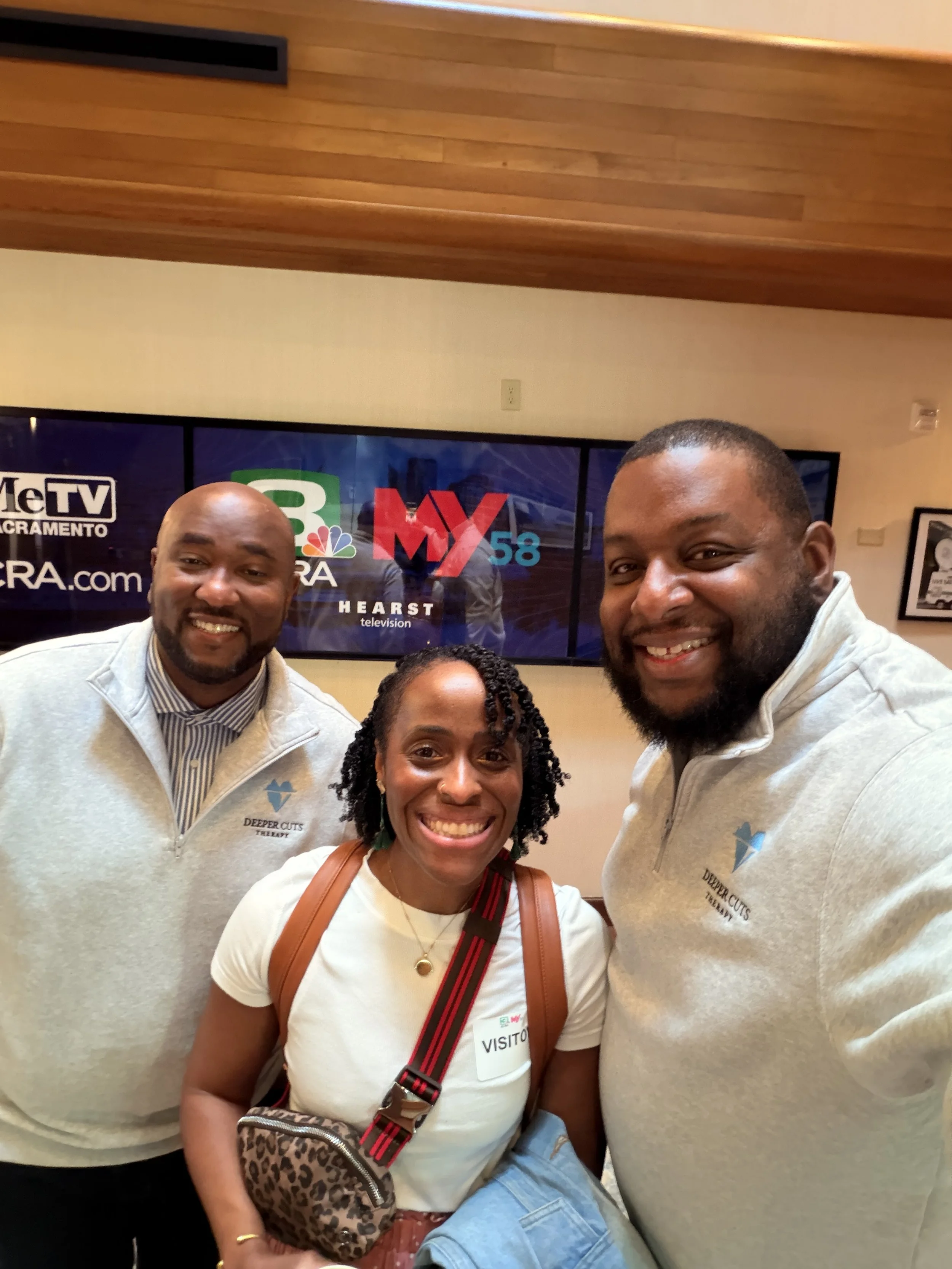 Four people smiling and taking a group photo in front of a television screen with news logos, including ABC, NBC, CBS, and others.