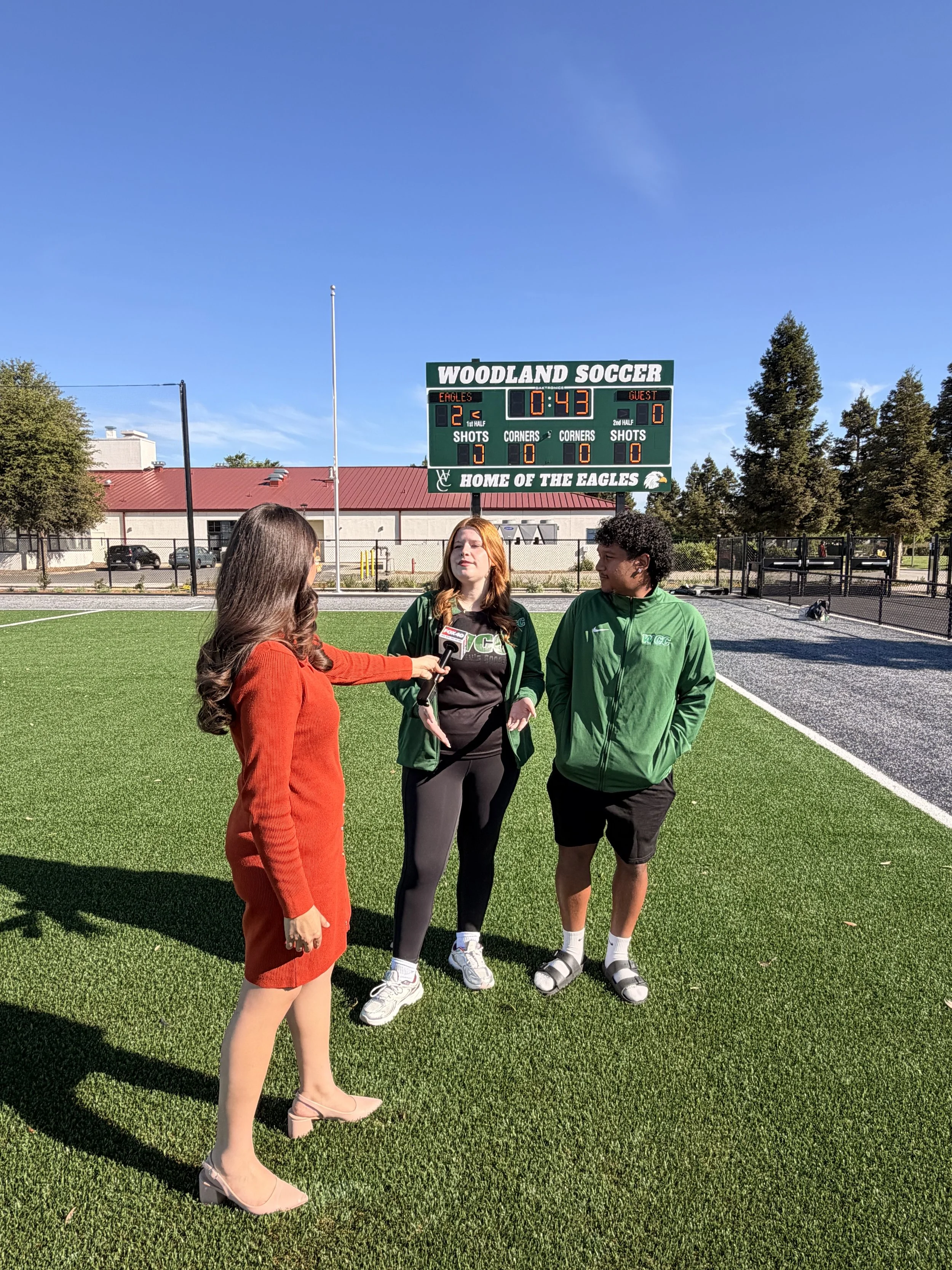Three teenagers are being interviewed on a soccer field near a scoreboard that reads 'Woodland Soccer' and shows the score as 2-0 with 43 seconds remaining in the game. The girl on the left is wearing a red dress, while the girl in the middle and boy