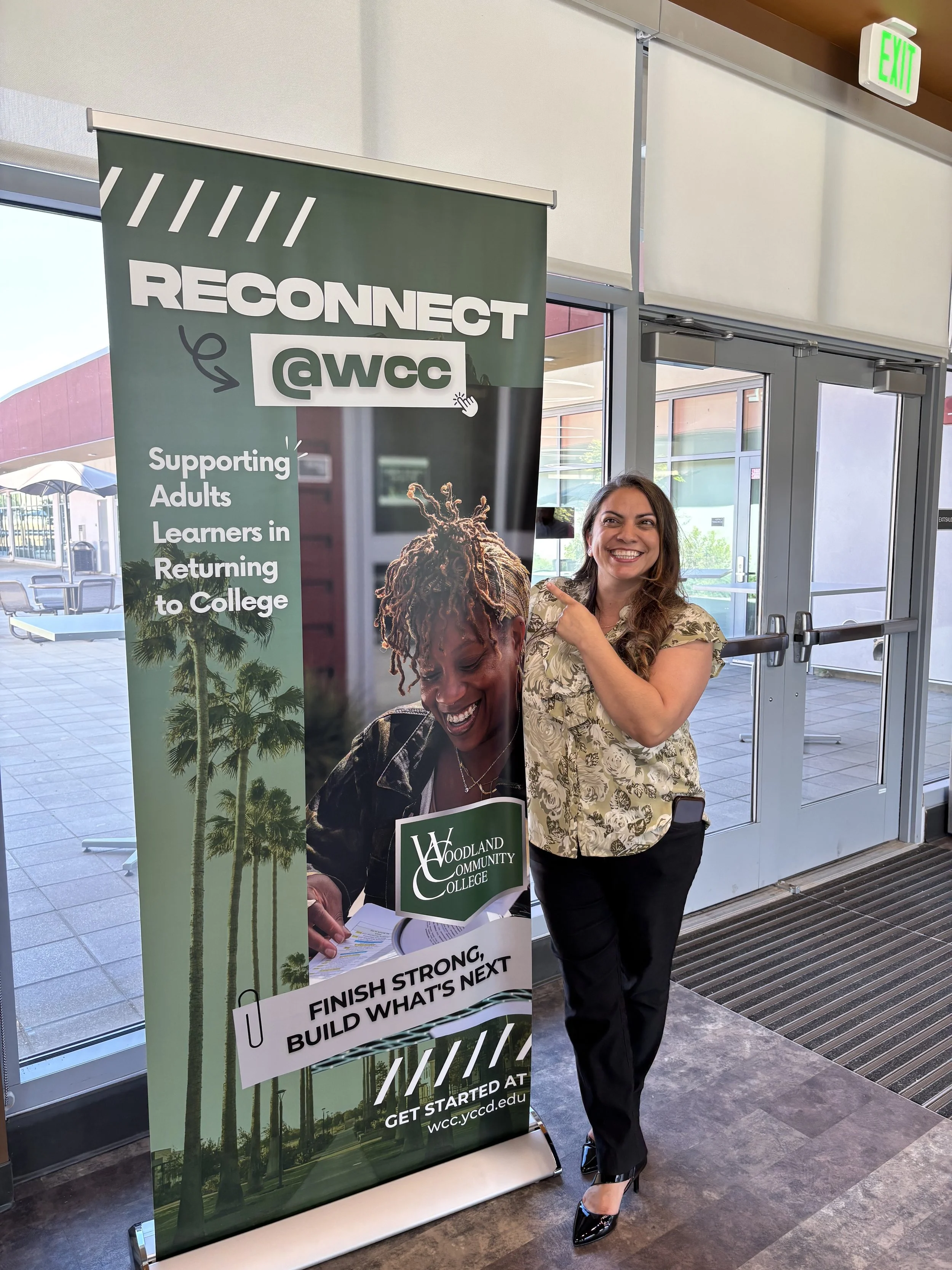 A woman with long brown hair and a floral blouse pointing to a banner for Woodland Community College, which supports adults, learners in returning to college, and has information about starting at the college.