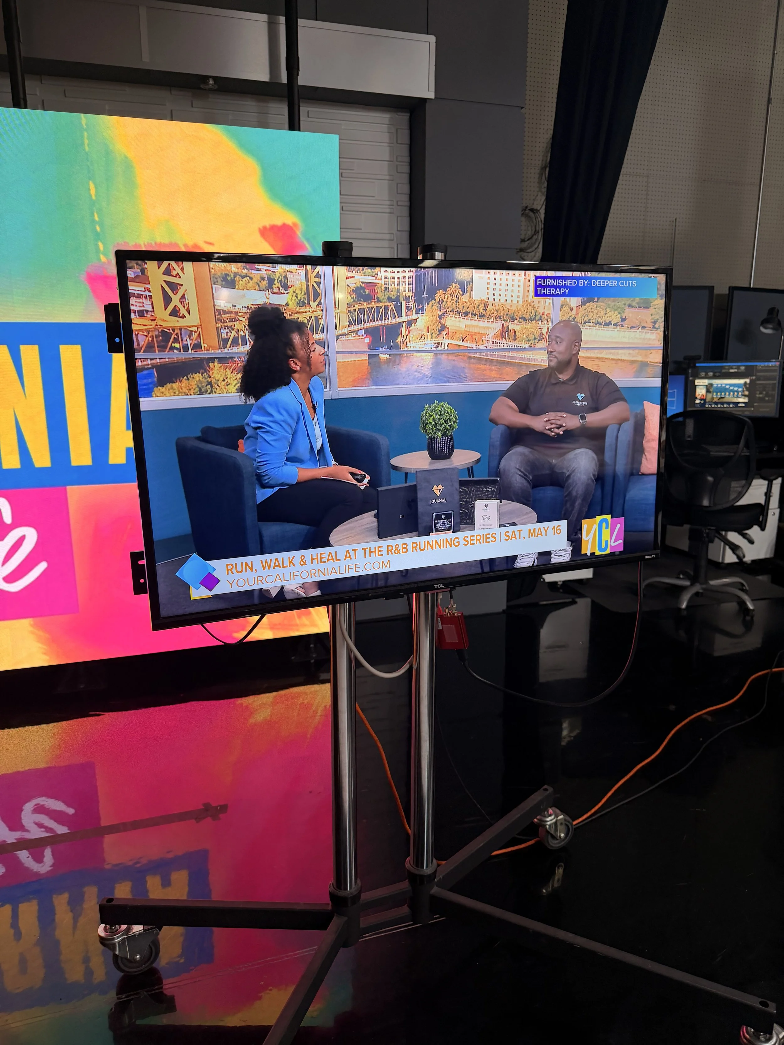Television screen showing a talk show with two people seated, a woman with curly hair in a blue blazer and a man in a black shirt, with a cityscape background. There is a banner at the bottom with event details and logos.