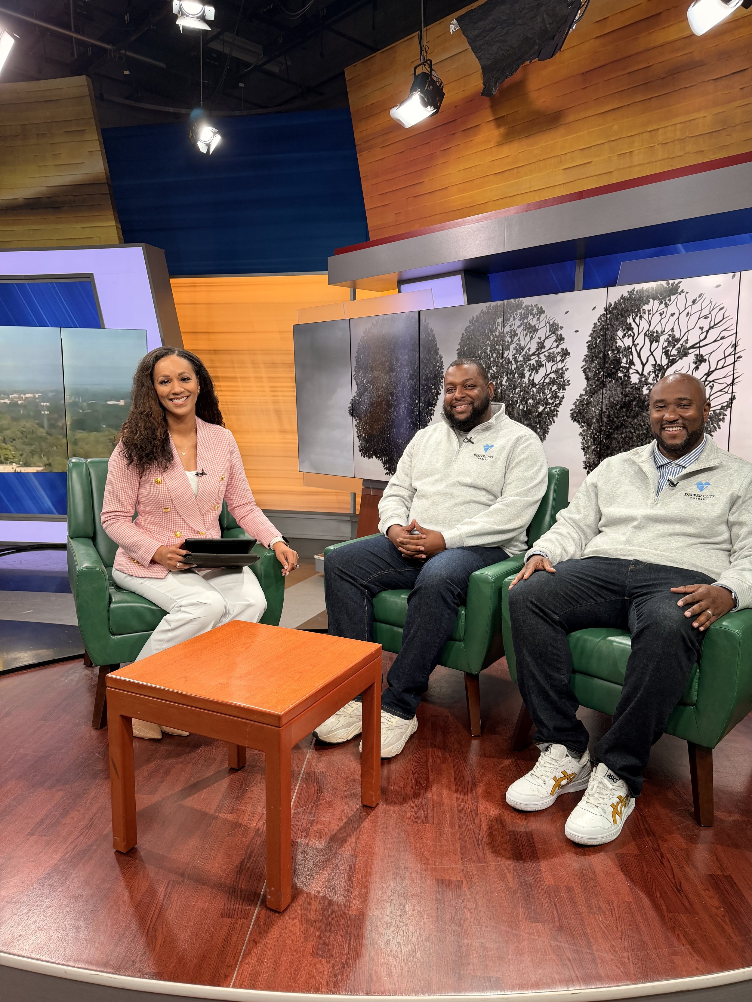 people sitting on green chairs in a television studio set with wooden and colored walls behind them. The woman on the left is smiling and holding a tablet, wearing a pink blazer. The three men on the right are smiling, wearing light gray sweatshirts 