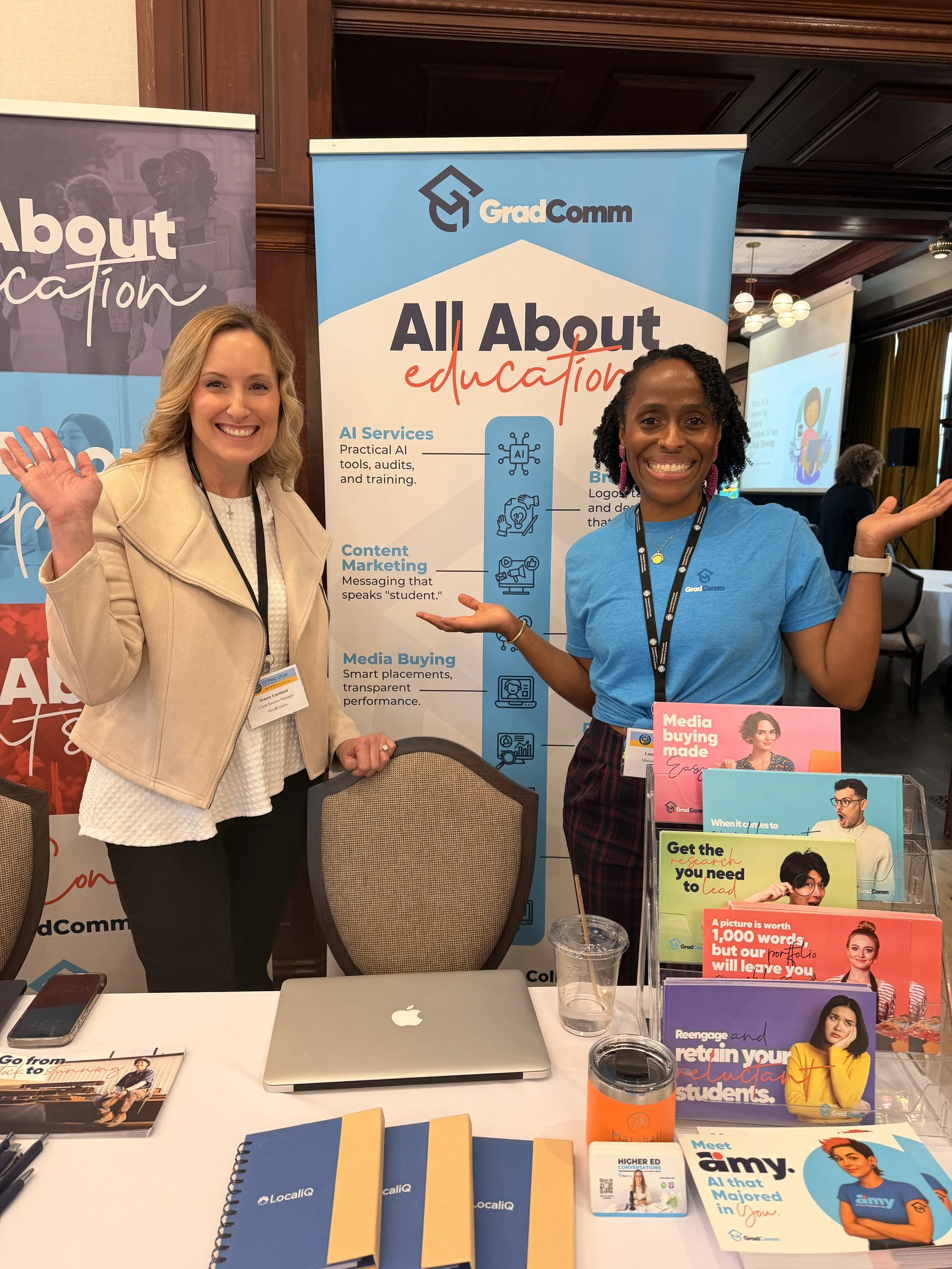 Two women standing behind a booth at a conference or expo, smiling and gesturing towards promotional materials and a banner behind them that reads 'All About Education' with icons and text about AI services, content marketing, and media buying.