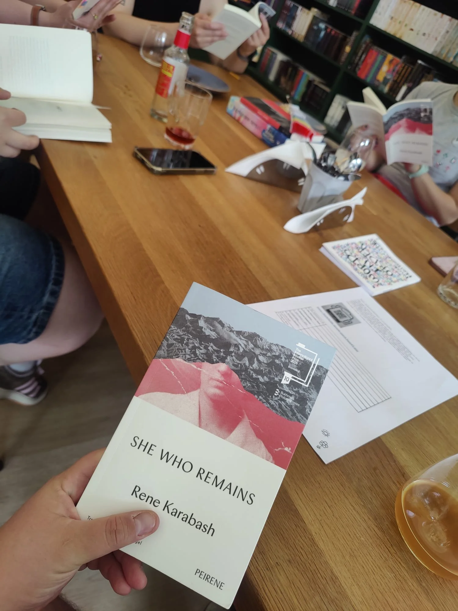 A person holding a book titled "She Who Remains" by Rene Karabash at a table with other books, notebooks, a smartphone, a bottle of alcohol, and a glass of wine. There are other people reading books in the background.