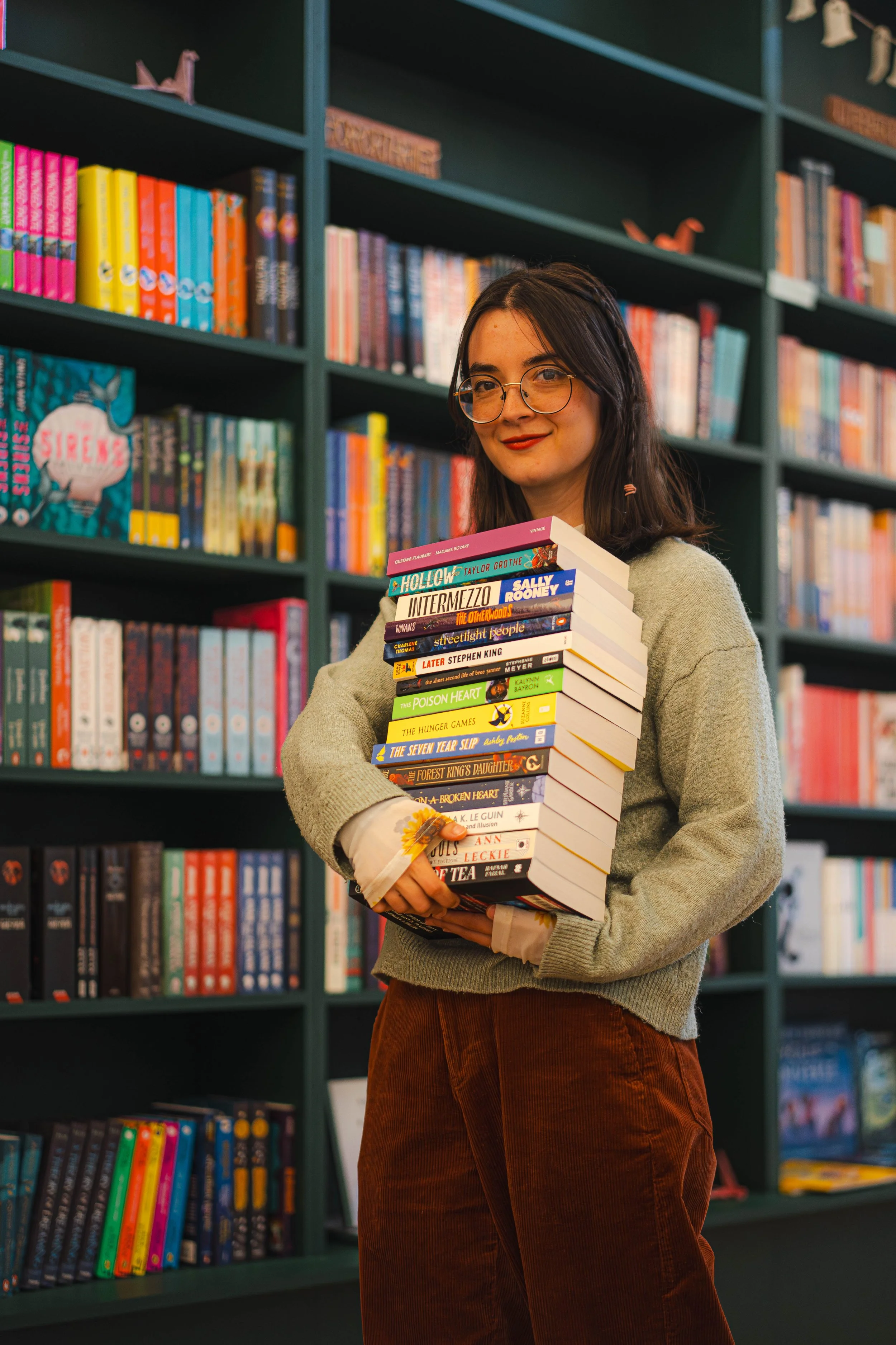 A woman with glasses and dark hair holding a stack of books in a bookstore.