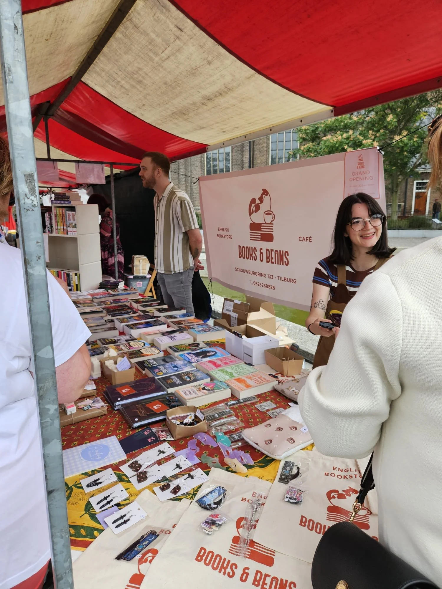 A booth at a book and coffee fair with people browsing books and merchandise, with a sign reading "Books & Beans" and details of a grand opening event in Tilburg.