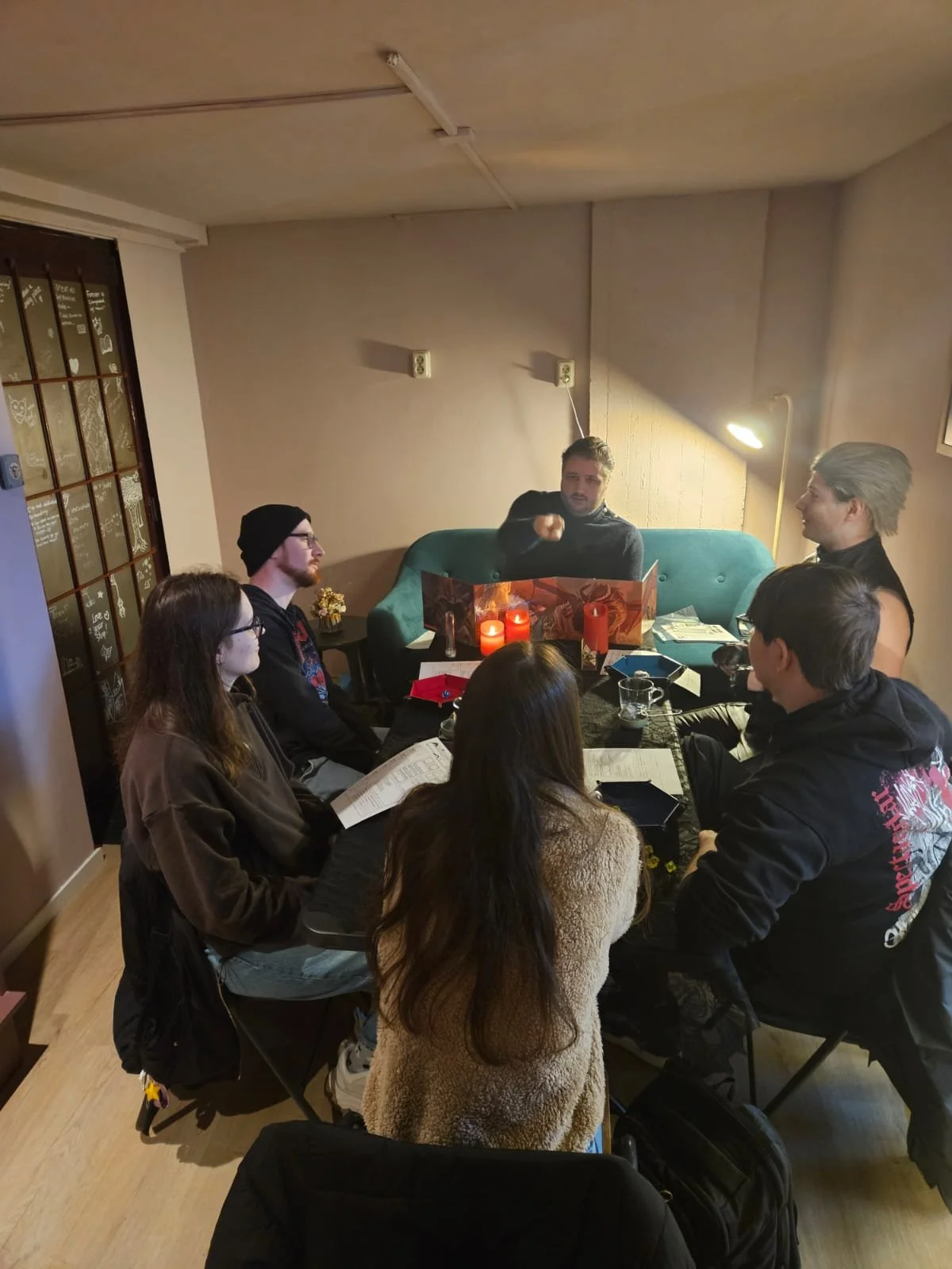 Group of seven people, four women and three men, gathered around a table in a cozy room. The man at the head is speaking and gesturing, with a book open in front of him. The others are listening, some with drinks and papers. Candles and a wall lamp p