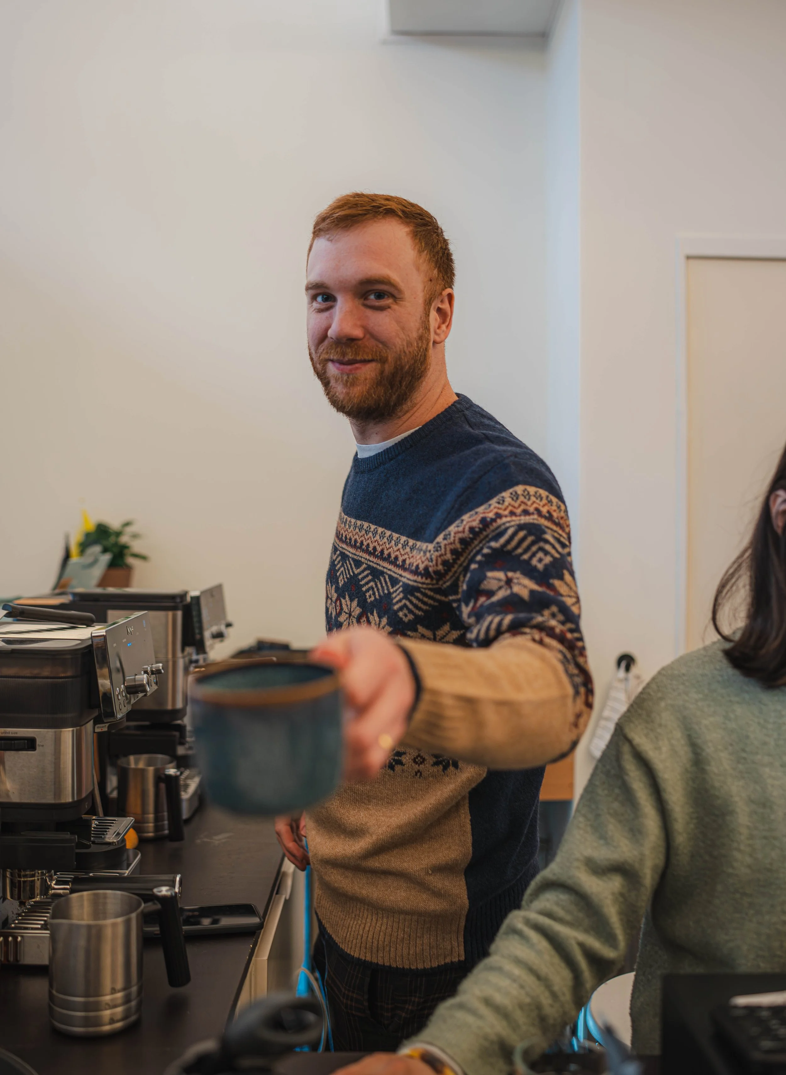 A man with red hair and a beard pouring coffee from a coffee machine in a kitchen or office break room, with another person partially visible nearby.
