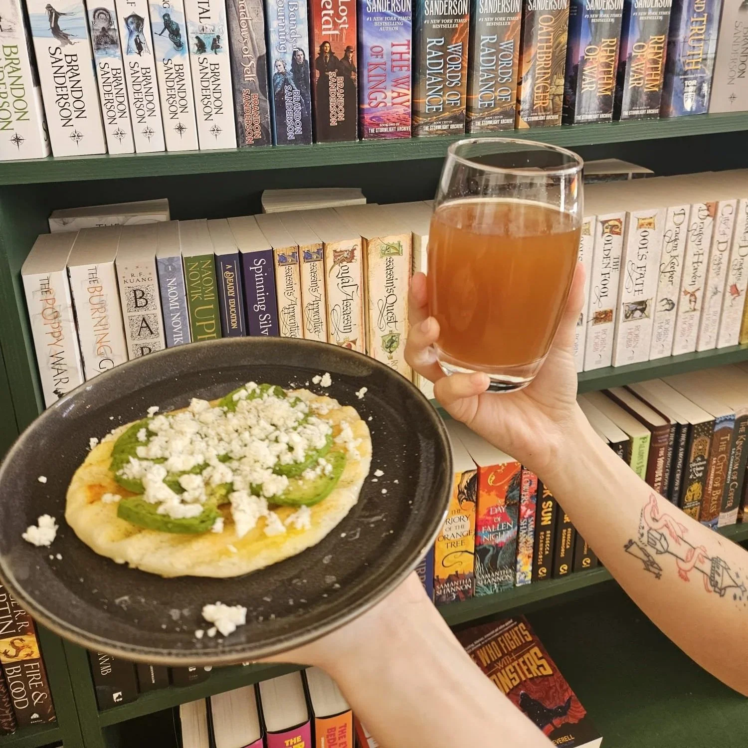 A hand holding a glass of rosé wine, another hand holding a black plate with a flatbread topped with green peppers and crumbled cheese, in front of a bookshelf filled with books.