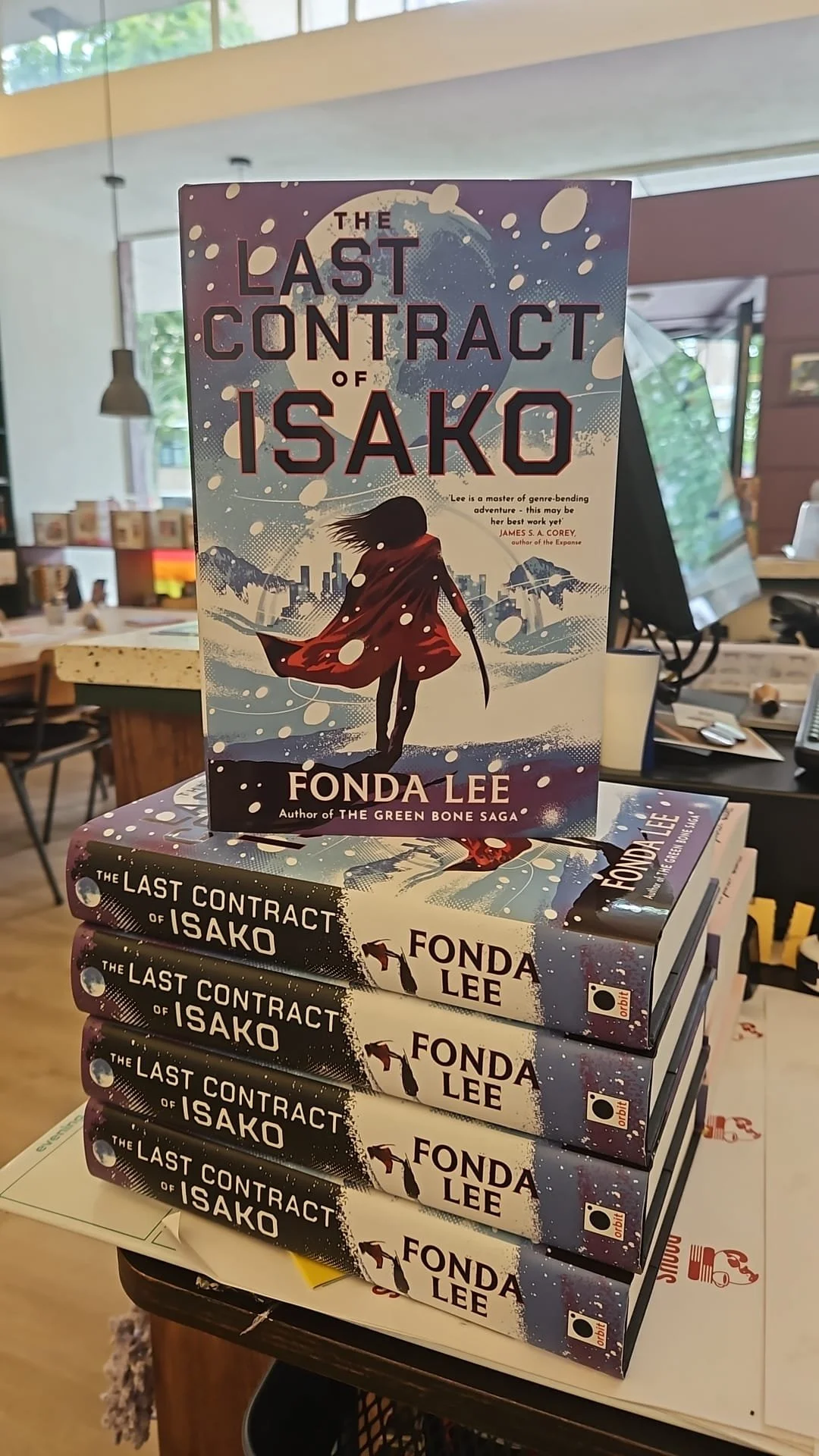 A stack of books titled "The Last Contract of Isako" by Fonda Lee on display in a bookstore. The front cover shows a silhouette of a girl with long hair, wearing a flowing cape, walking in a snowy landscape with a city skyline and mountains in the background.
