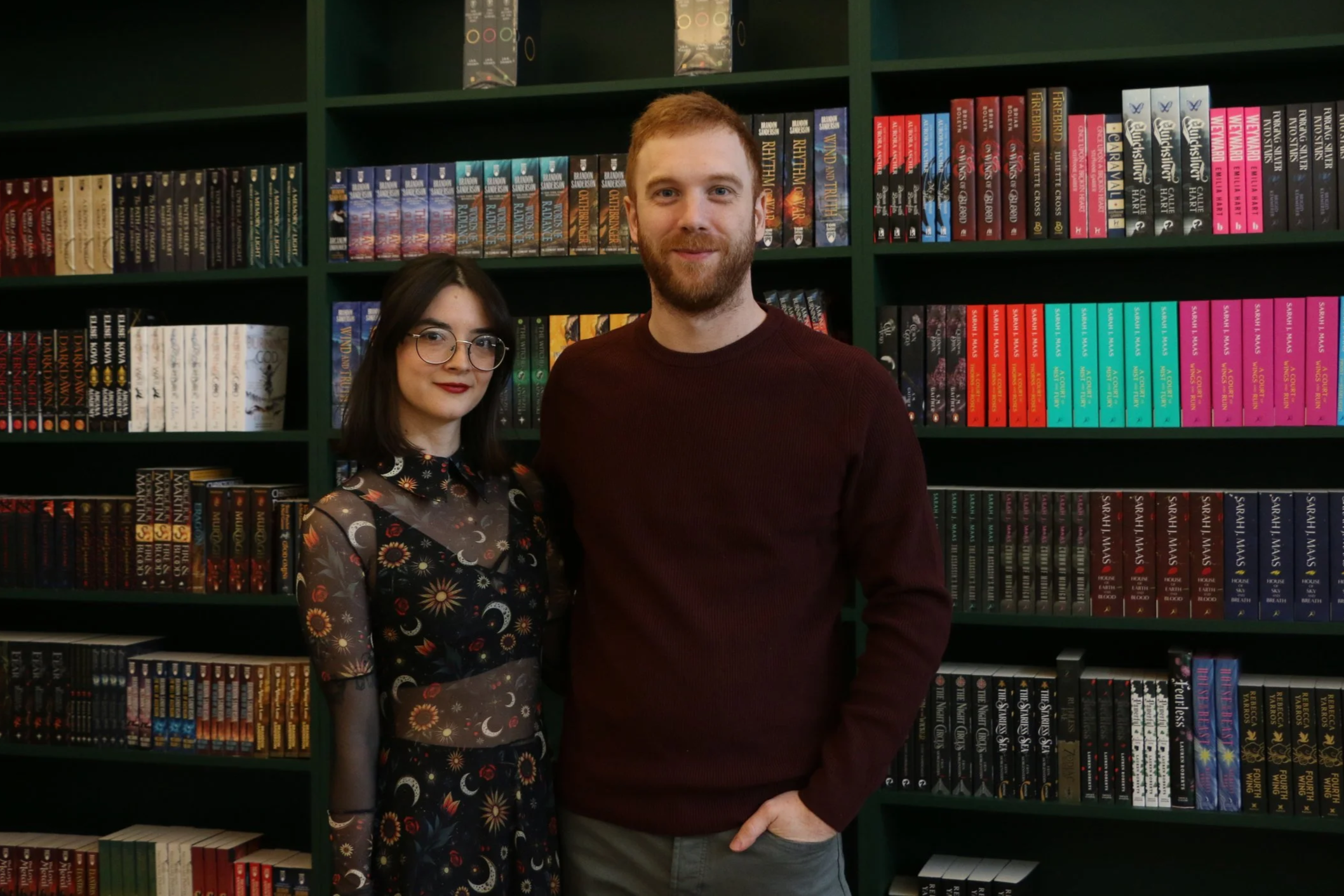 Two people, a woman with dark hair and glasses wearing a colorful patterned dress, and a man with red hair and a beard wearing a dark red sweater and gray pants, standing in front of bookshelves filled with colorful books.