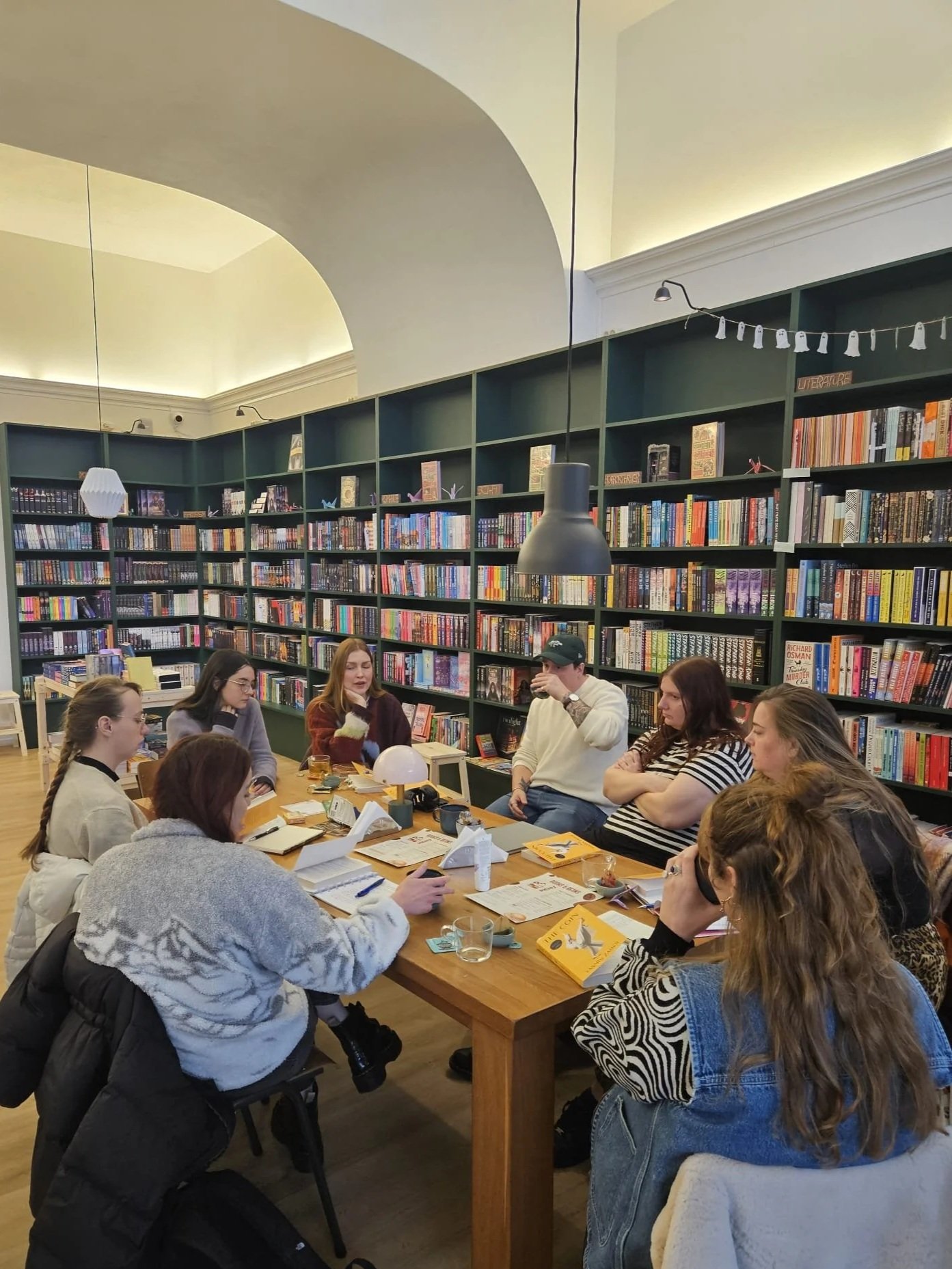 Group of people sitting around a wooden table in a bookstore or library, with shelves full of books in the background, engaging in a discussion or book club meeting.