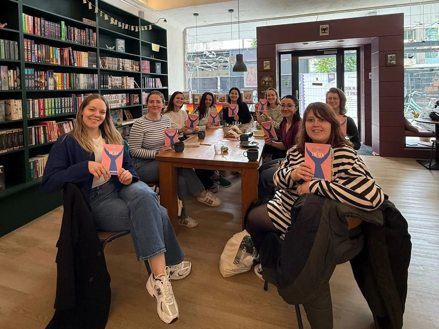 A group of women sitting around a wooden table in a bookstore cafe, each holding a pink book with a blue whale illustration on the cover.