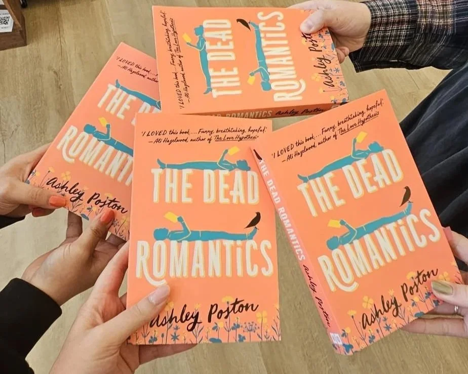 Books titled "The Dead Romantics" by Ashley Poston are held and fanned out in a group. The cover features an orange background with illustrations of a person lying down, a bird, and flowers.