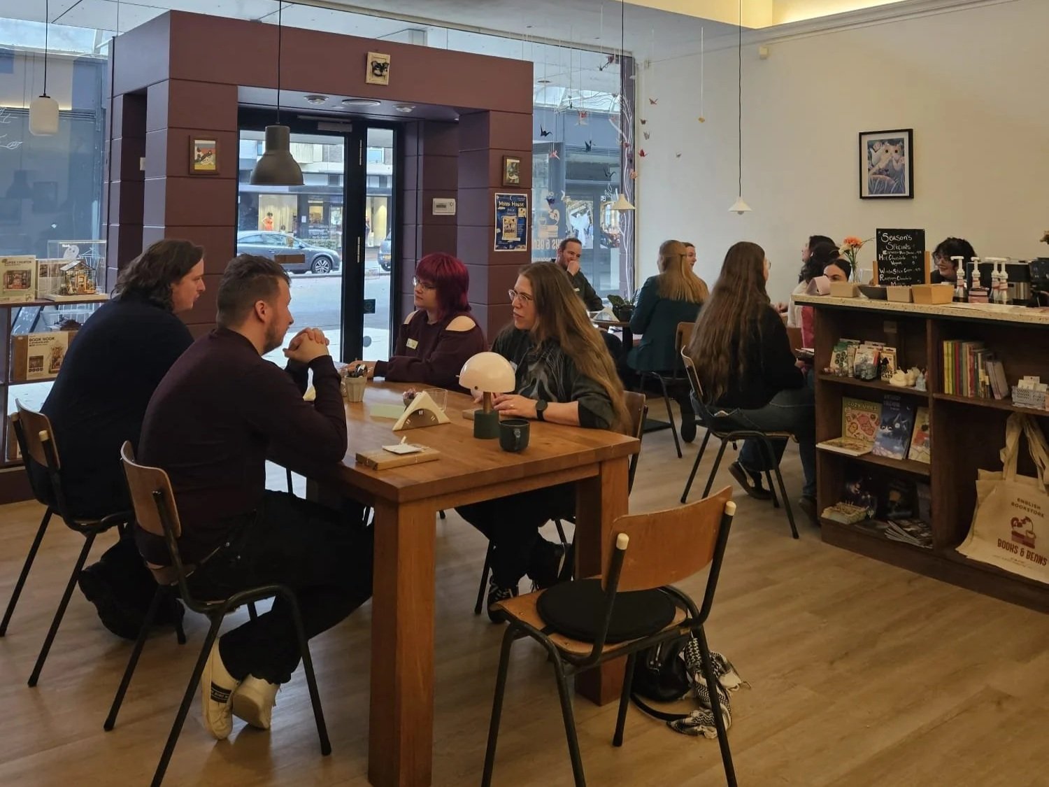 People sitting and chatting at tables in a cozy coffee shop with bookshelves, artwork, and a large window letting in natural light.