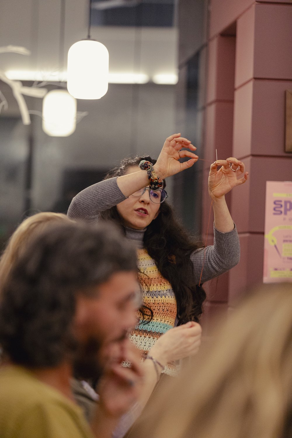 A woman with long dark hair, wearing glasses and a rainbow-colored knitted vest, demonstrates how to knit or crochet with a needle. She is indoors, with warm lighting and pink-colored walls. There are other people in the background.