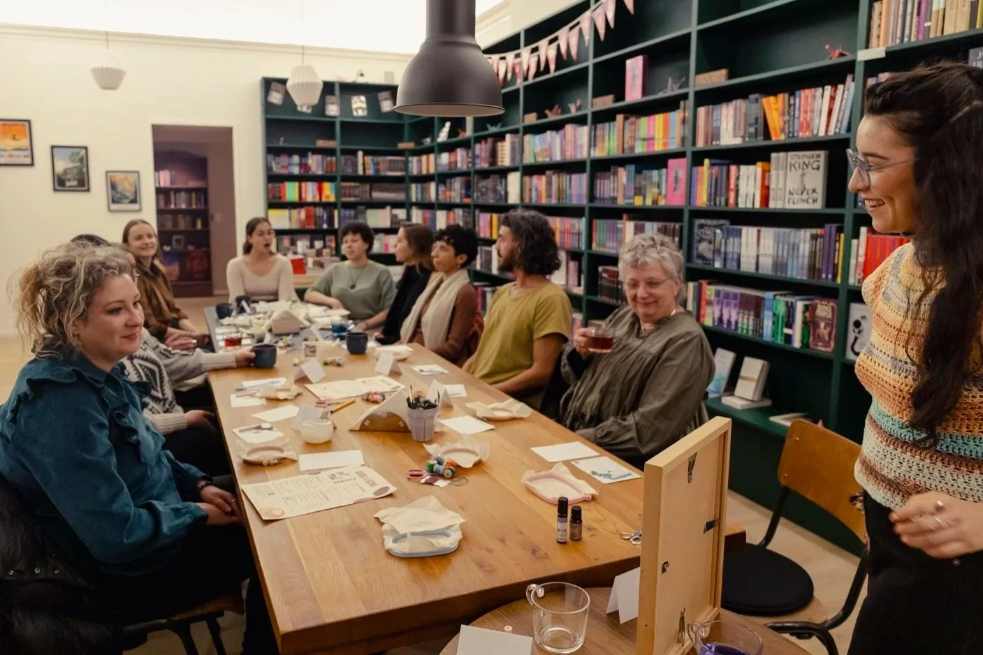 Group of women sitting around a wooden table in a bookstore, engaging in a meeting or workshop, with various items like papers, pens, and drinks on the table, and bookshelves filled with books in the background.