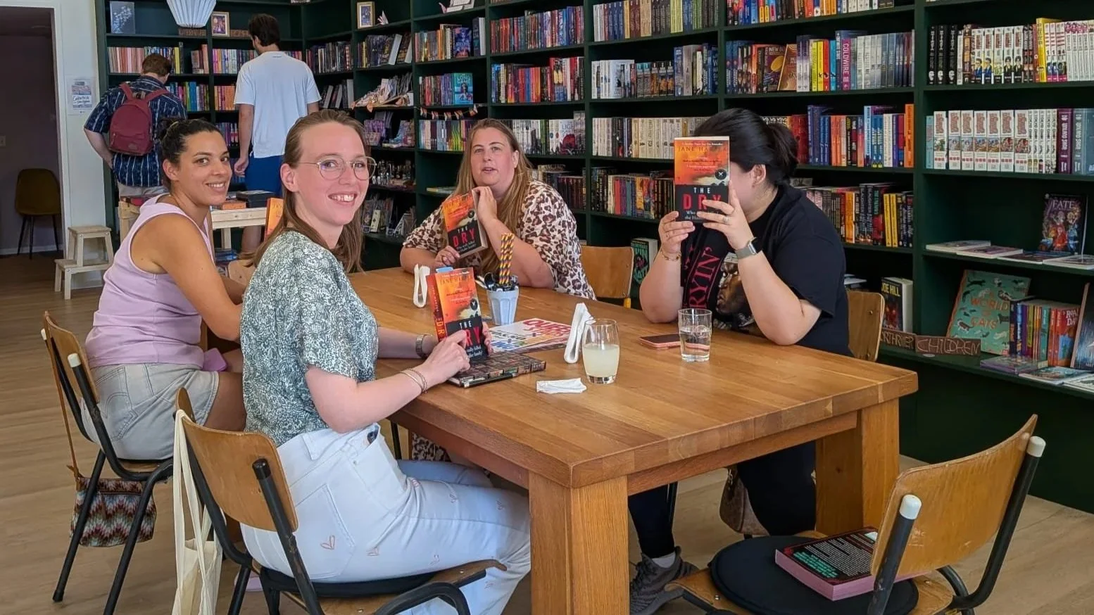 Four women sitting around a wooden table in a bookstore, holding books titled 'The Dory'. There are shelves of books in the background and two people in the distance browsing.