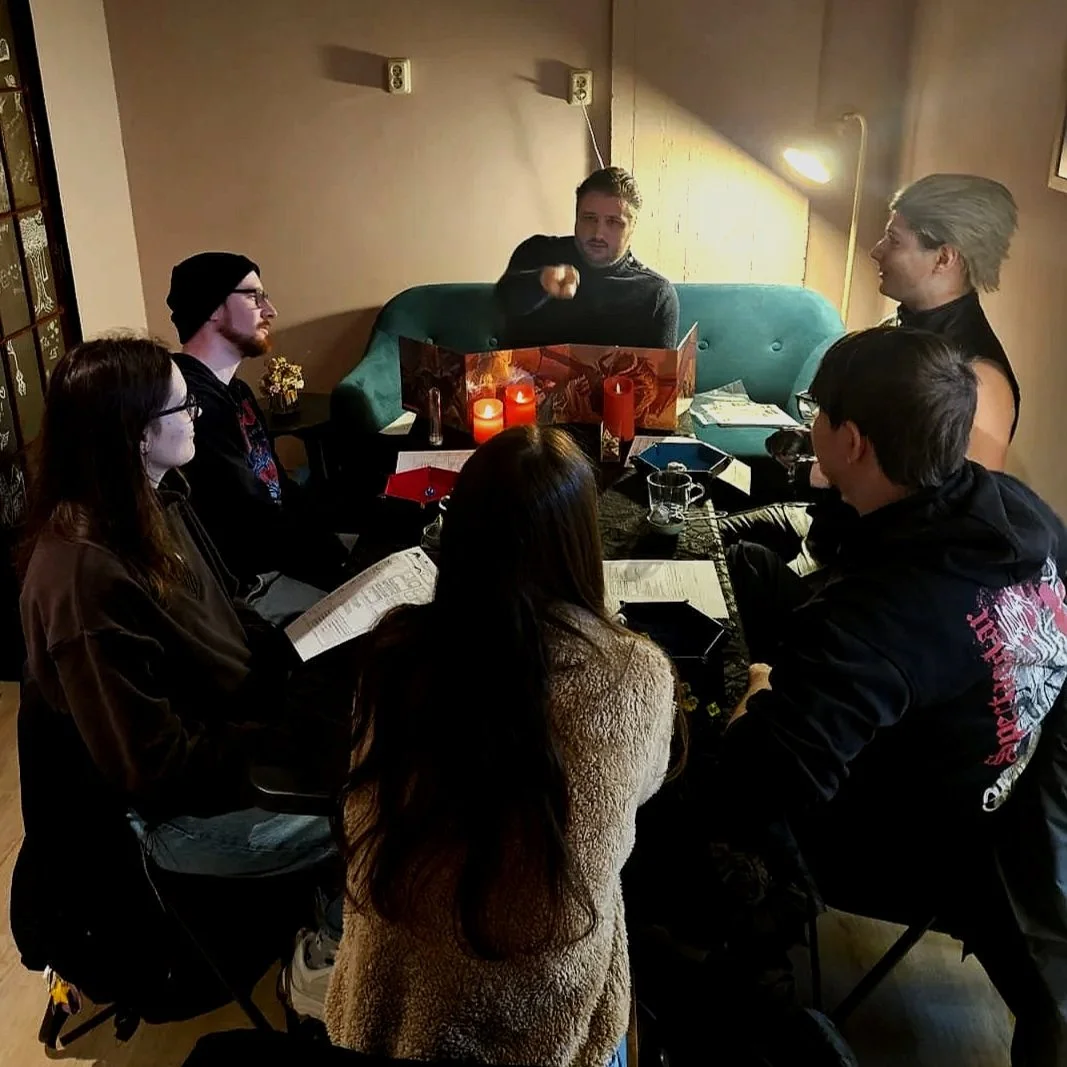 Group of six people sitting around a table playing Dungeons and Dragons, with a game master leading the session. Candles and drinks are on the table in a cozy, dimly lit room.