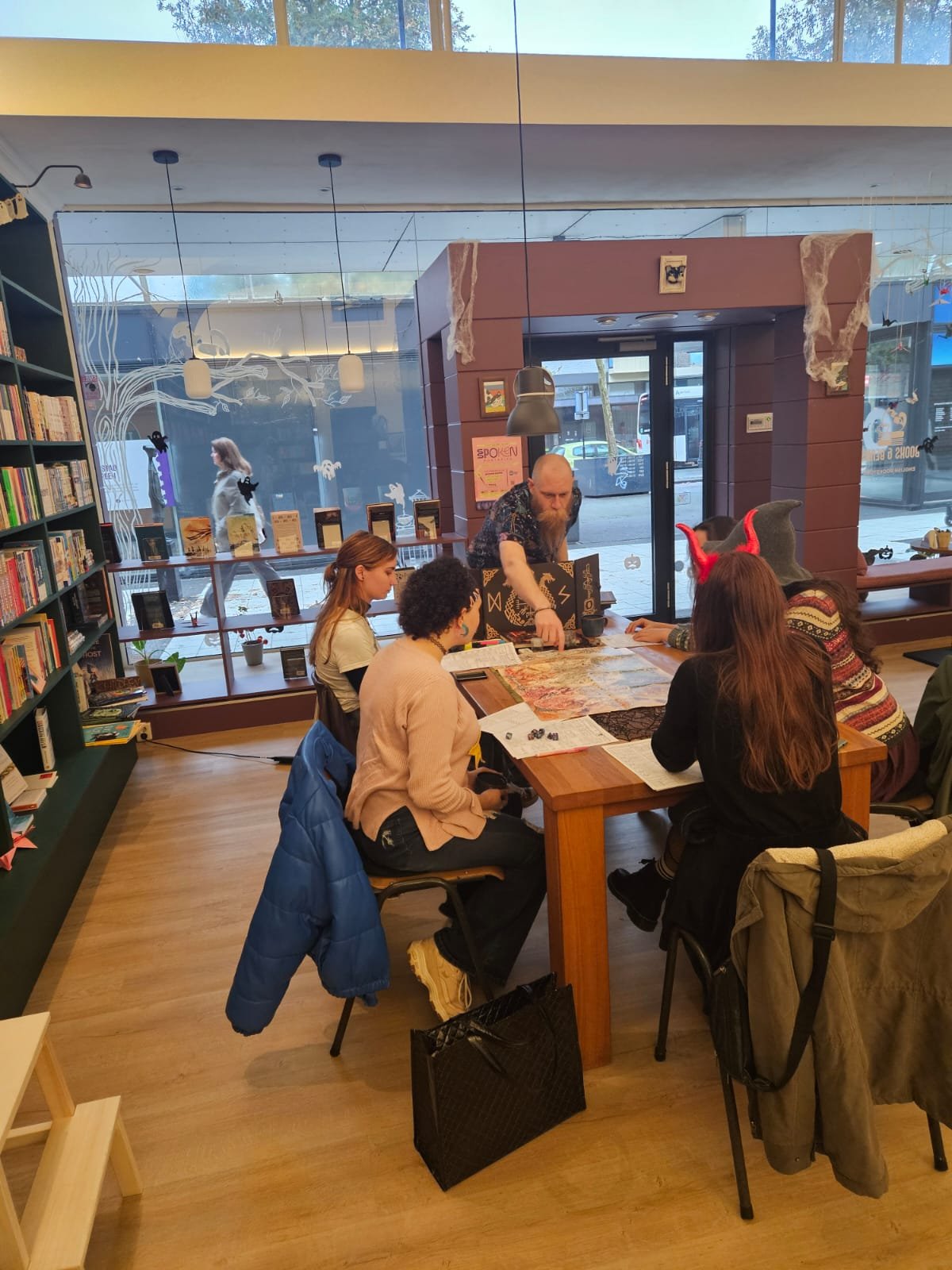 People sitting around a table in a bookstore, playing a tabletop game, with a person dressed as a demon with red horns leading the game.