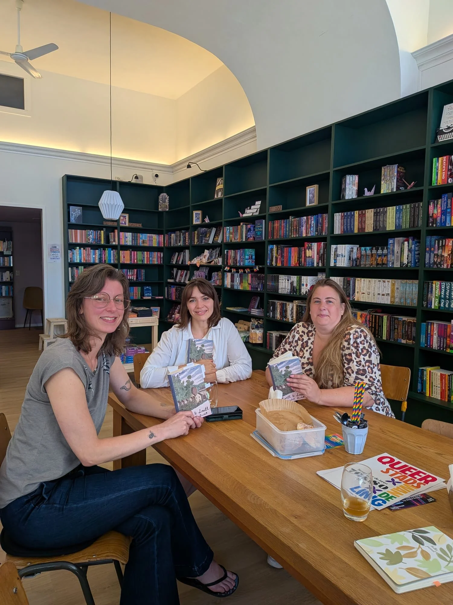 Three women sitting at a wooden table in a bookstore or library, holding books, with bookshelves filled with colorful books in the background.