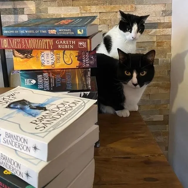 Two black and white cats sitting on a wooden table next to a stack of books. The books include titles by Brandon Sanderson, Michael Livingston, Gareth Rubin, and C.S. Lewis, with a brick wall in the background.
