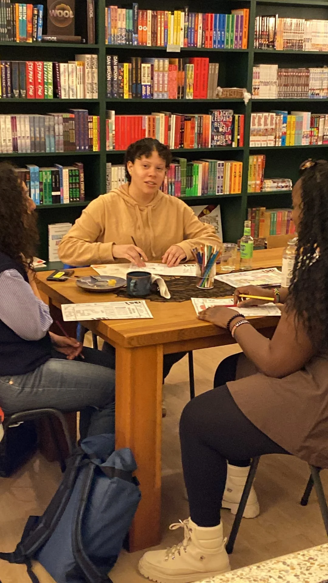 A group of four people sitting around a wooden table in a bookstore or library, engaging in a discussion or activity, with bookshelves filled with colorful books in the background.