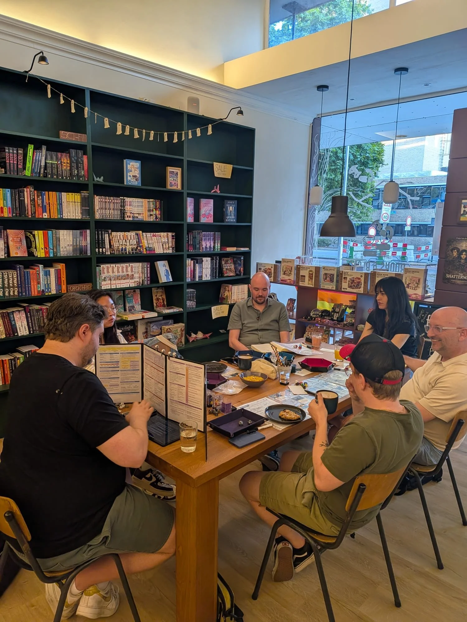 A group of people playing tabletop role-playing game at a wooden table in a bookstore or library with bookshelves, large windows, and modern lighting.