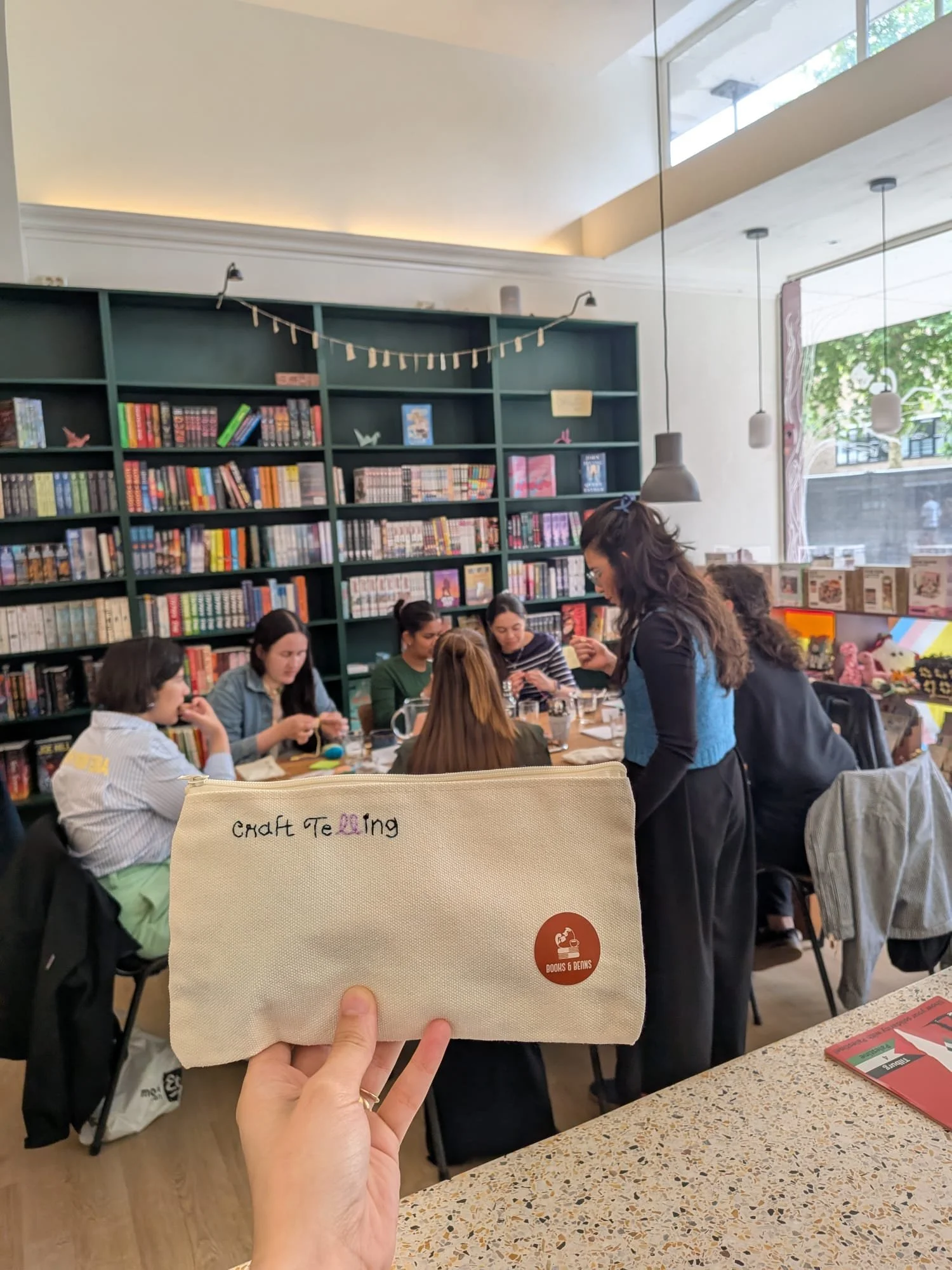 A group of people sitting at a table in a bookstore or library, with a bookshelf filled with colorful books and decorative items in the background. One person is standing and interacting with others, and the photo is taken from inside the store. A ha