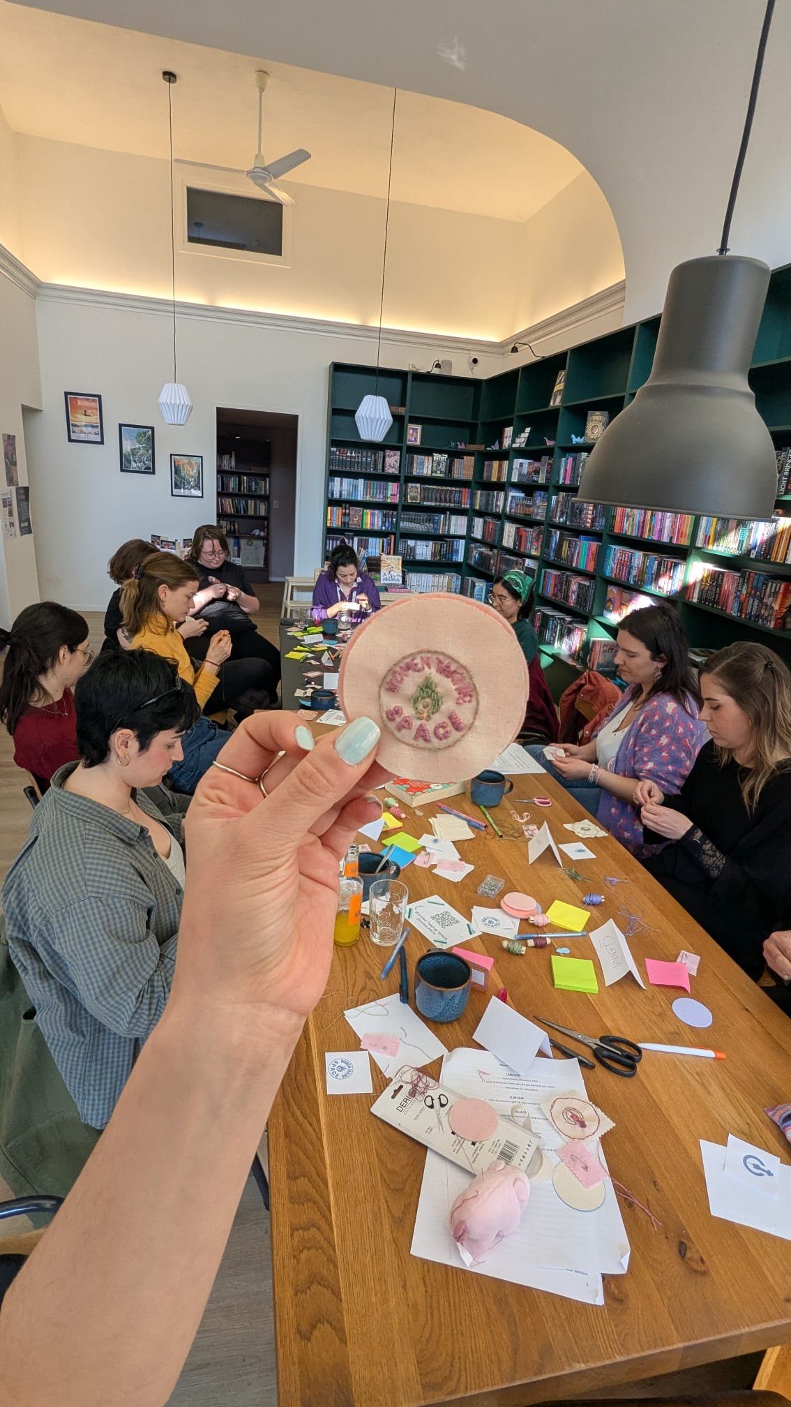 A group of people gathered around a large table in a library, engaged in arts and crafts activities, with a hand holding a fabric patch with embroidered text in the foreground.