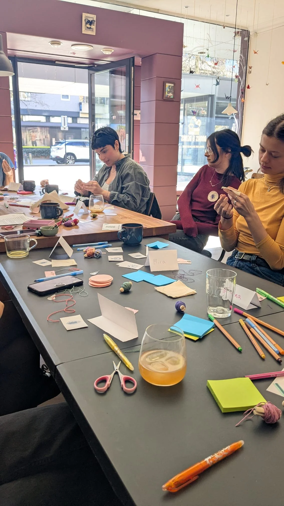People sitting at a table engaging in crafting activities in a brightly lit room with large windows. The table is cluttered with colorful paper, markers, scissors, yarn, and cups.