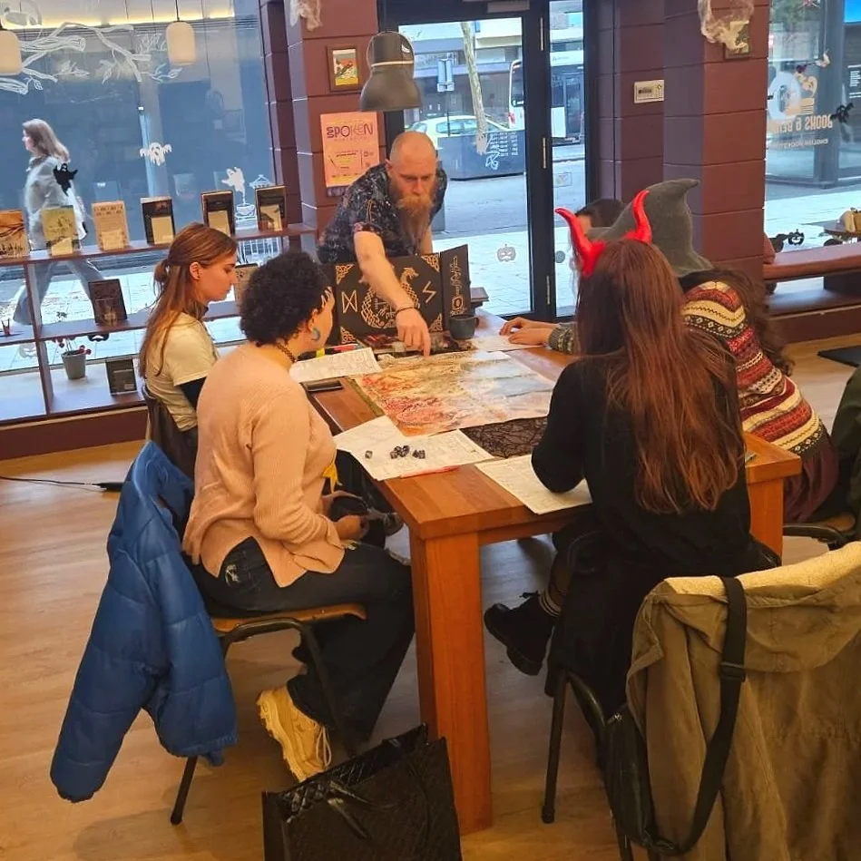 People gathered around a wooden table inside a cafe playing a tabletop game. Some are wearing costumes or accessories like devil horns and a witch hat. There are books and game pieces on the table, and the cafe has large windows and a brick wall.