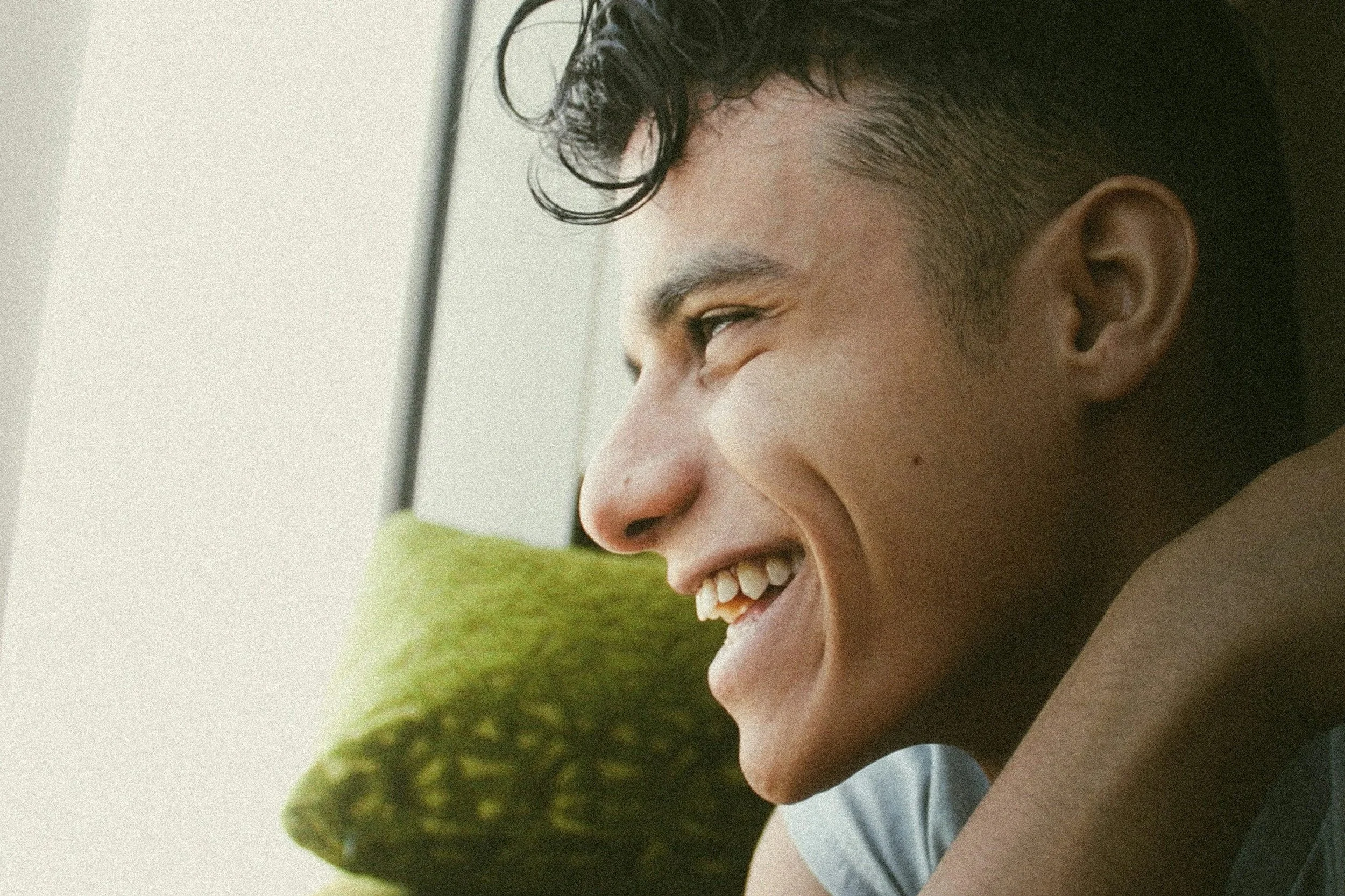 A young man with dark, curly hair, smiling and looking to the left. He has a light to medium skin tone and is sitting in front of a green textured pillow.