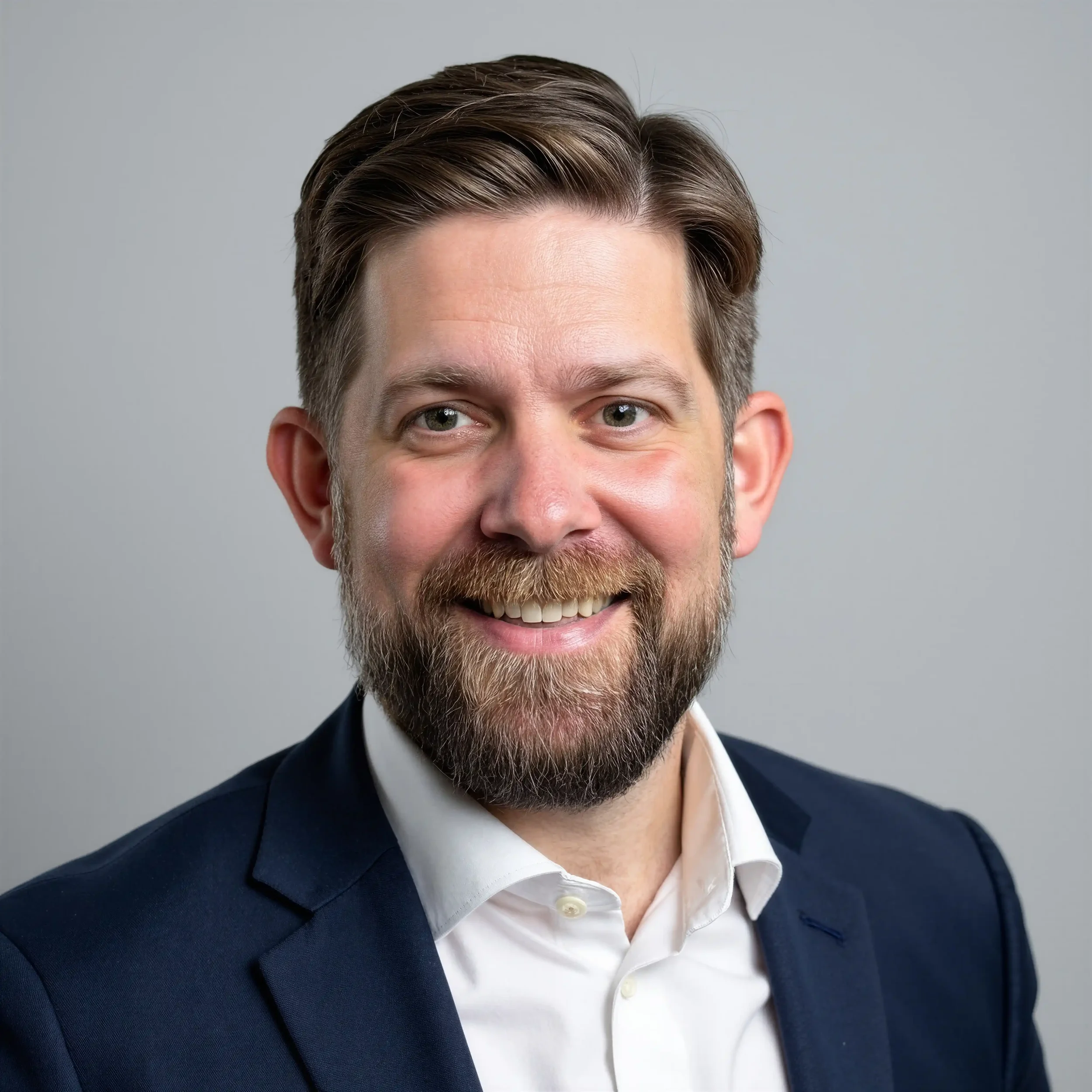 Close-up of Andy smiling with a well-groomed beard and neatly styled brown hair, wearing a dark suit jacket over a white shirt against a plain gray background.