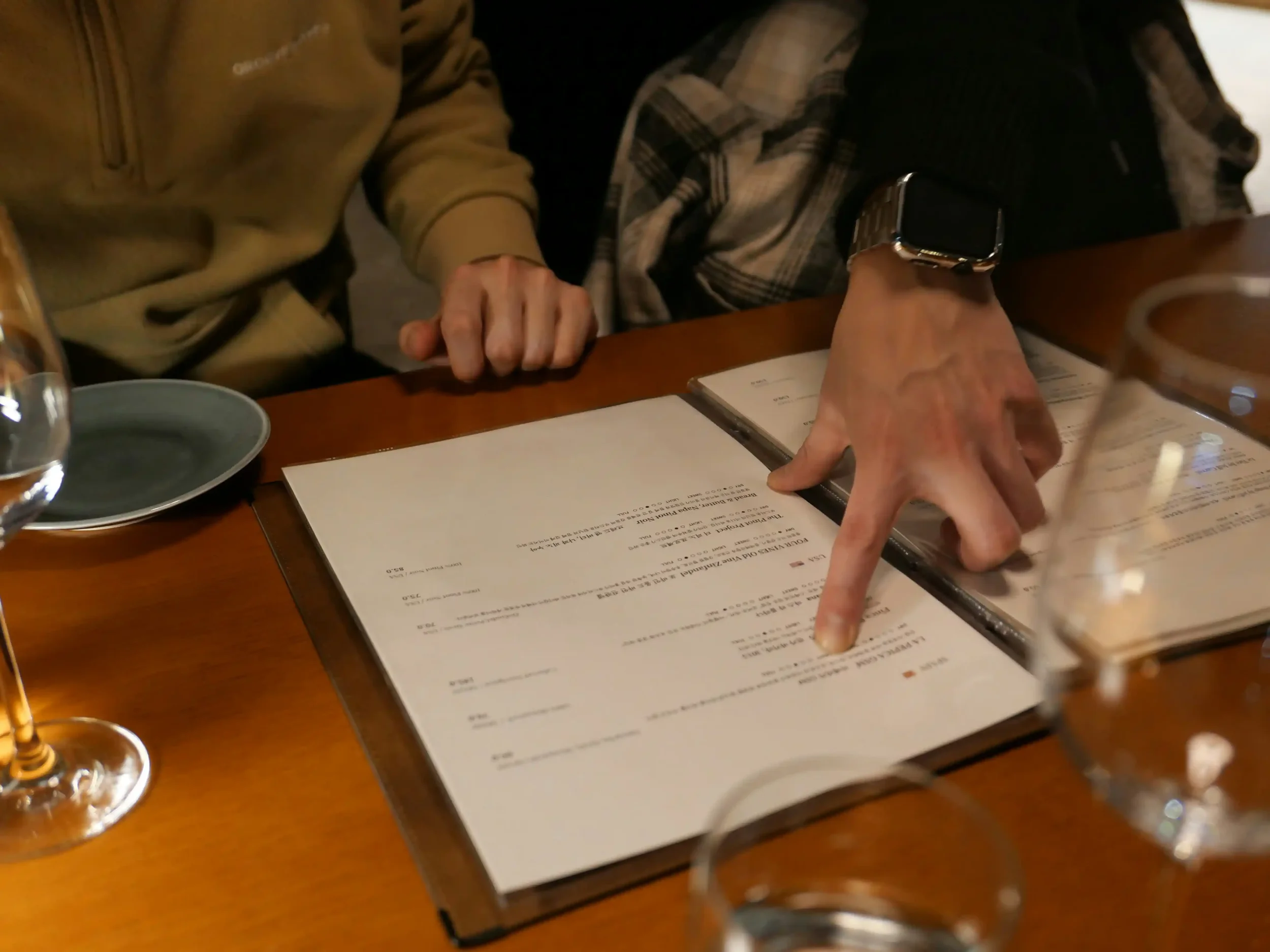 A person wearing a smartwatch points at a restaurant menu on a table, with another person's hand resting nearby and some glassware visible.