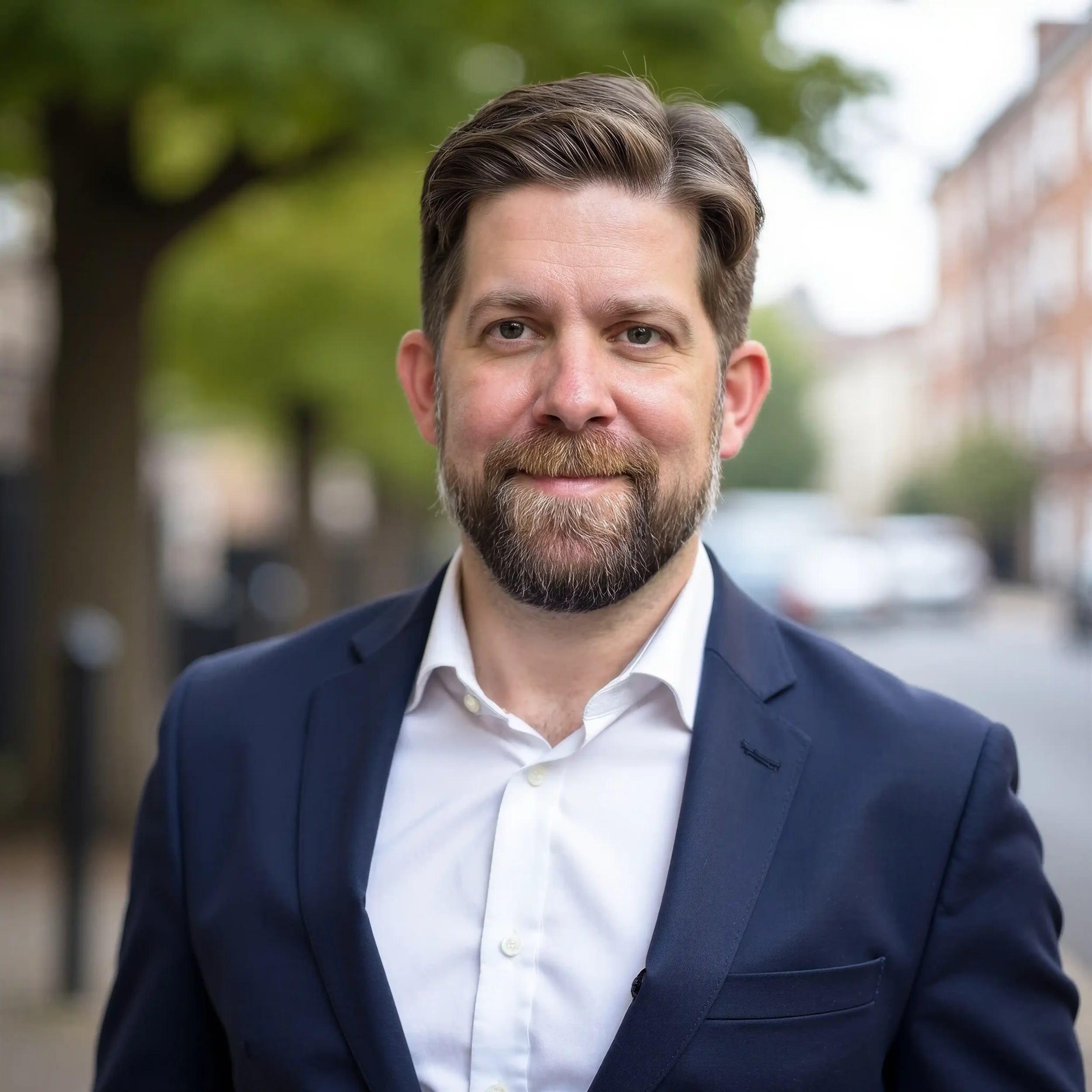 Close-up of Andy dressed in a dark blue suit and white shirt, with well-groomed hair and facial hair, standing outdoors with blurred trees and buildings in the background.