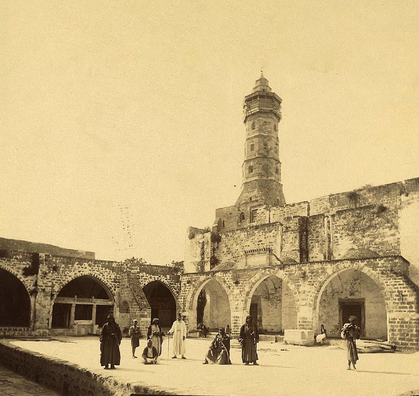 Historical sepia photograph of a mosque of Gaza with a tall minaret, stone arches, and several people in traditional clothing standing and walking in the courtyard.