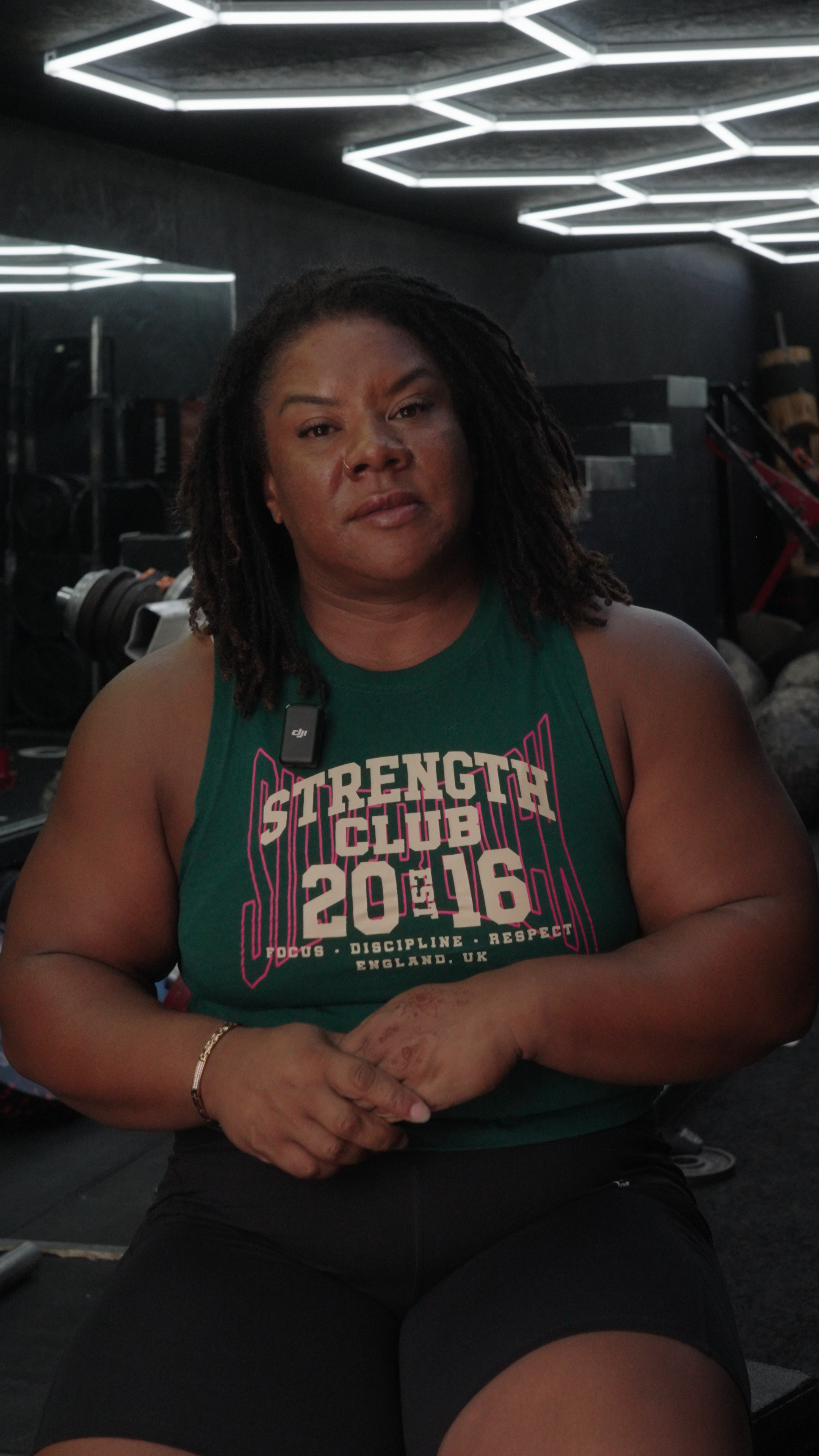 A woman with dark skin and dreadlocks sitting in a gym, wearing a green tank top with the text 'Strength Club 2016' and black shorts, with workout equipment visible in the background.