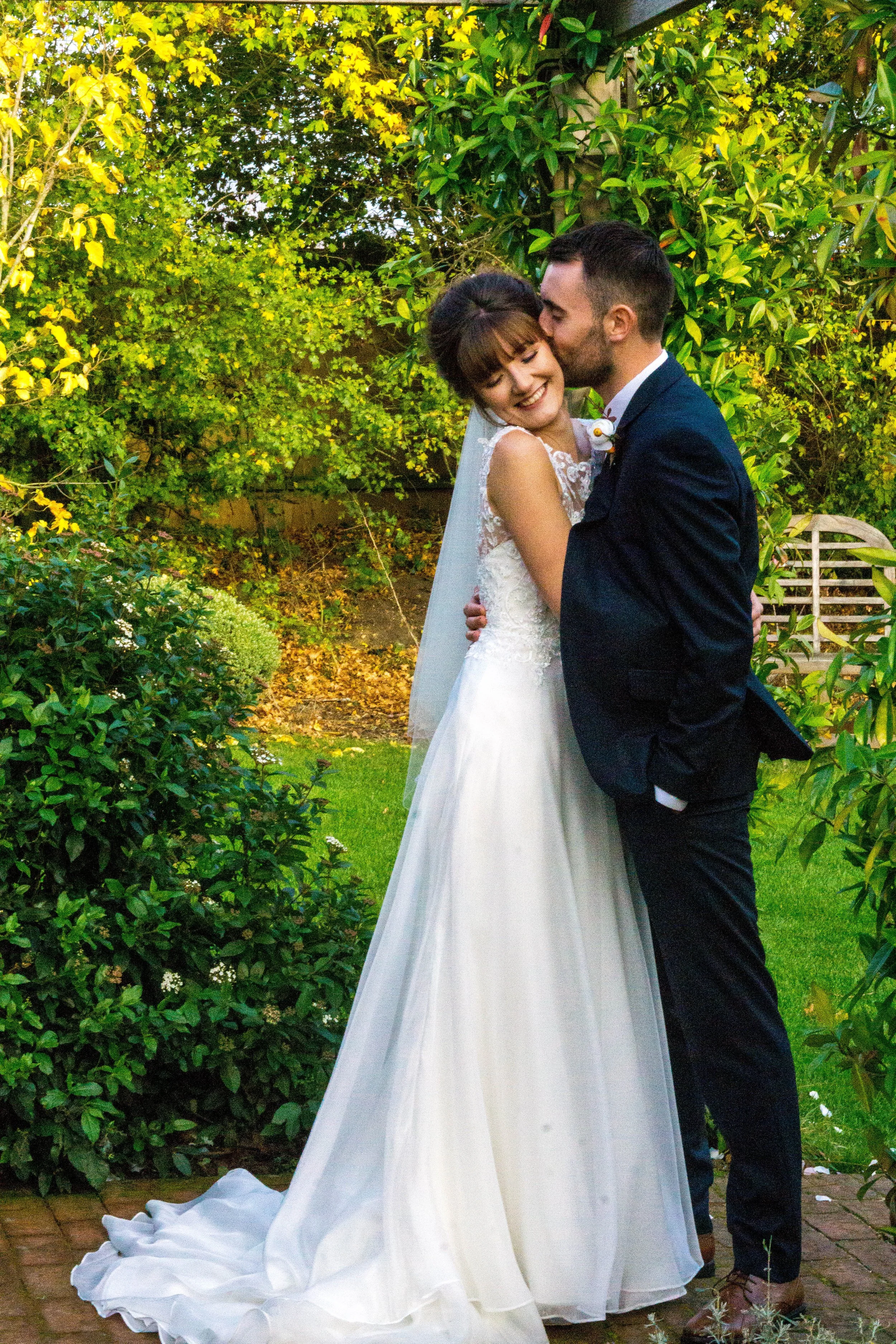 A bride and groom embrace outdoors in a garden, the groom kissing the bride on her temple. The bride is wearing a white wedding gown with lace details and the groom is dressed in a dark suit. The background is lush with greenery and yellow flowers.