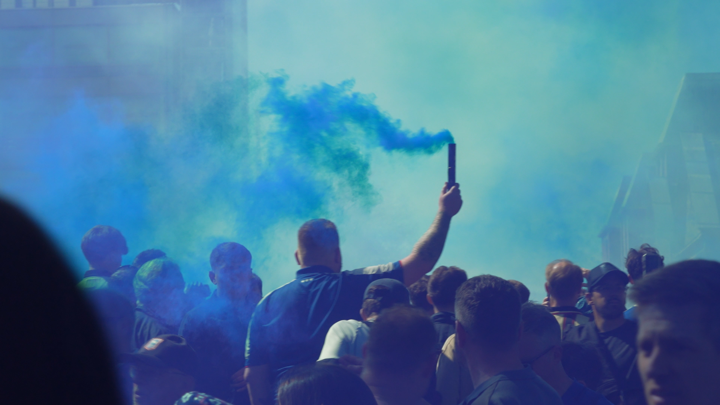Crowd of people at a public event with colored smoke and a person holding a smoke flare in the air.