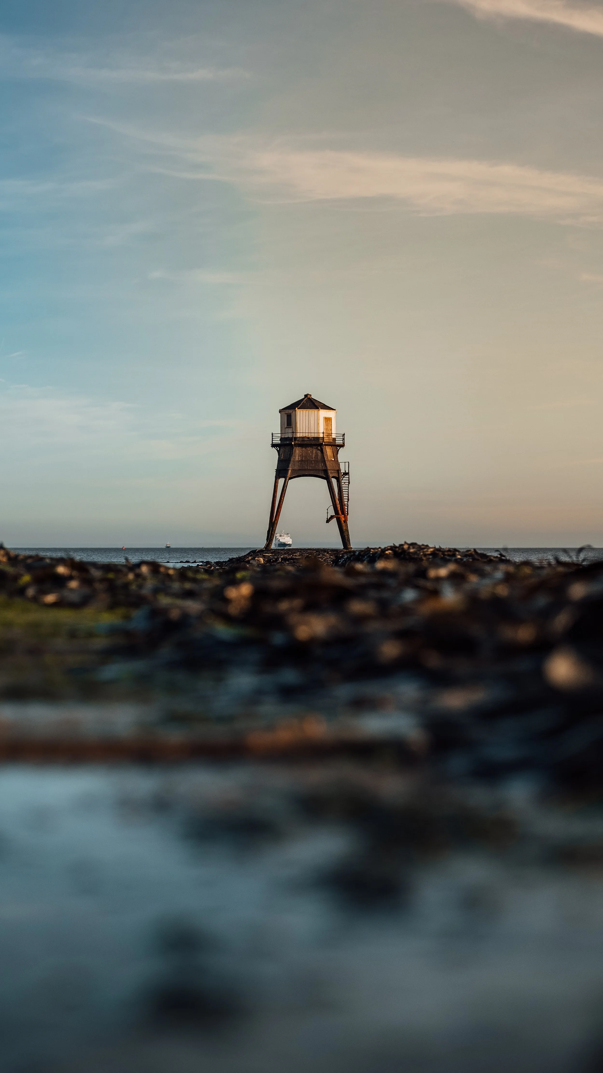 A small lighthouse stands on a rocky shoreline with the ocean in the background, under a cloudy sky during dusk or dawn.