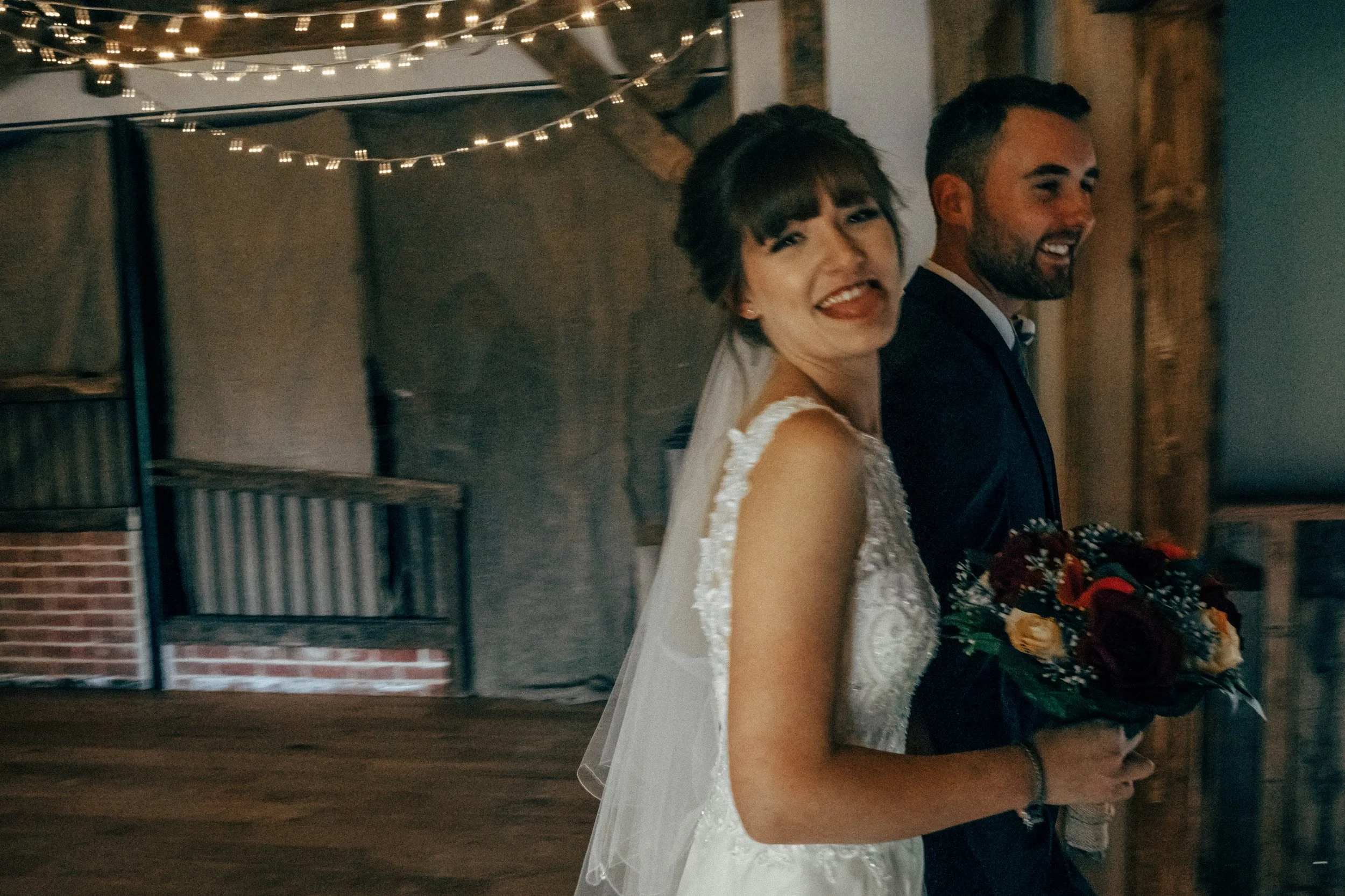 A bride and groom smiling at their wedding reception, standing close together against a rustic wooden wall with string lights overhead.