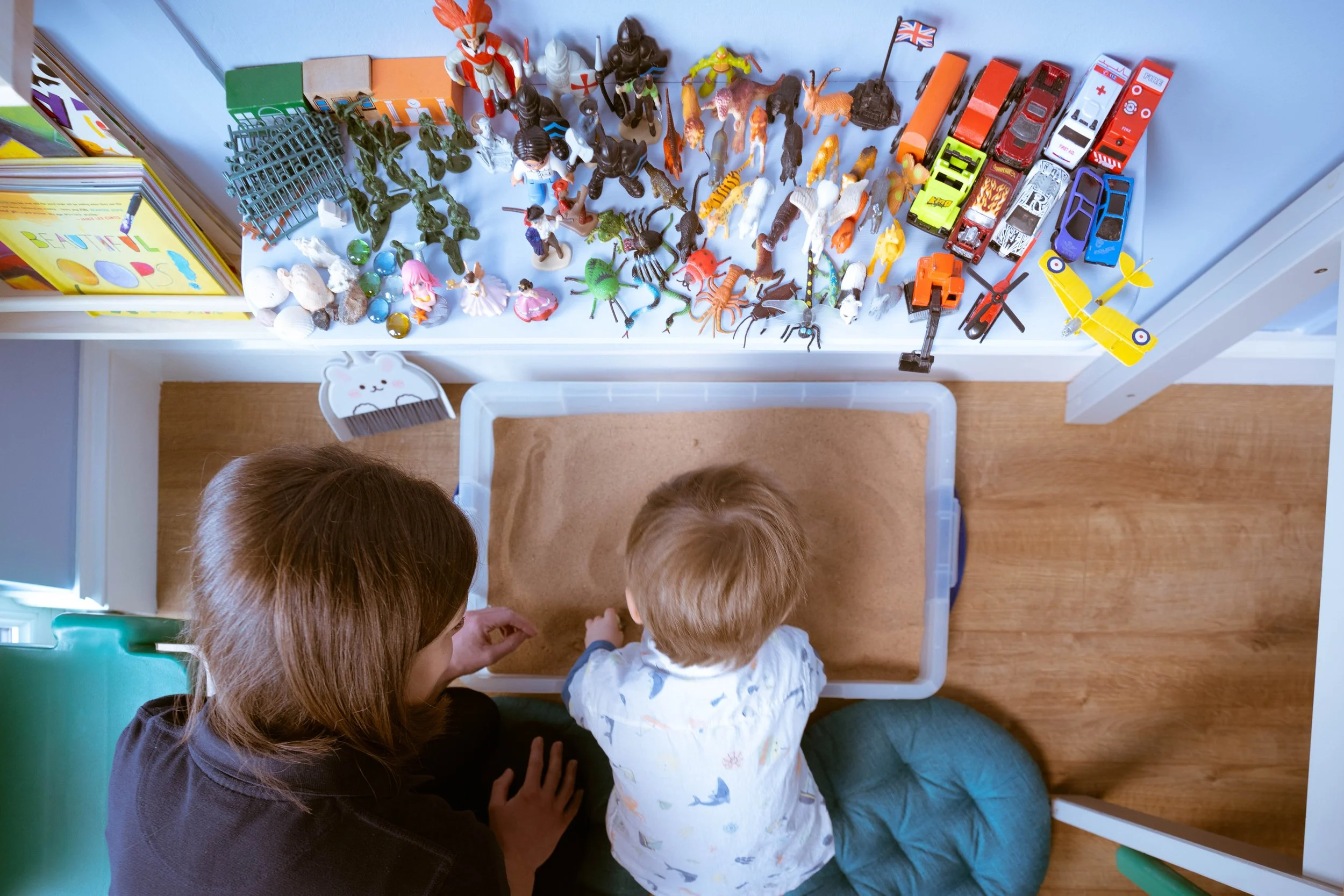 A woman and two children sitting on the floor, looking at a bookshelf with toy figurines, animals, and cars on top.