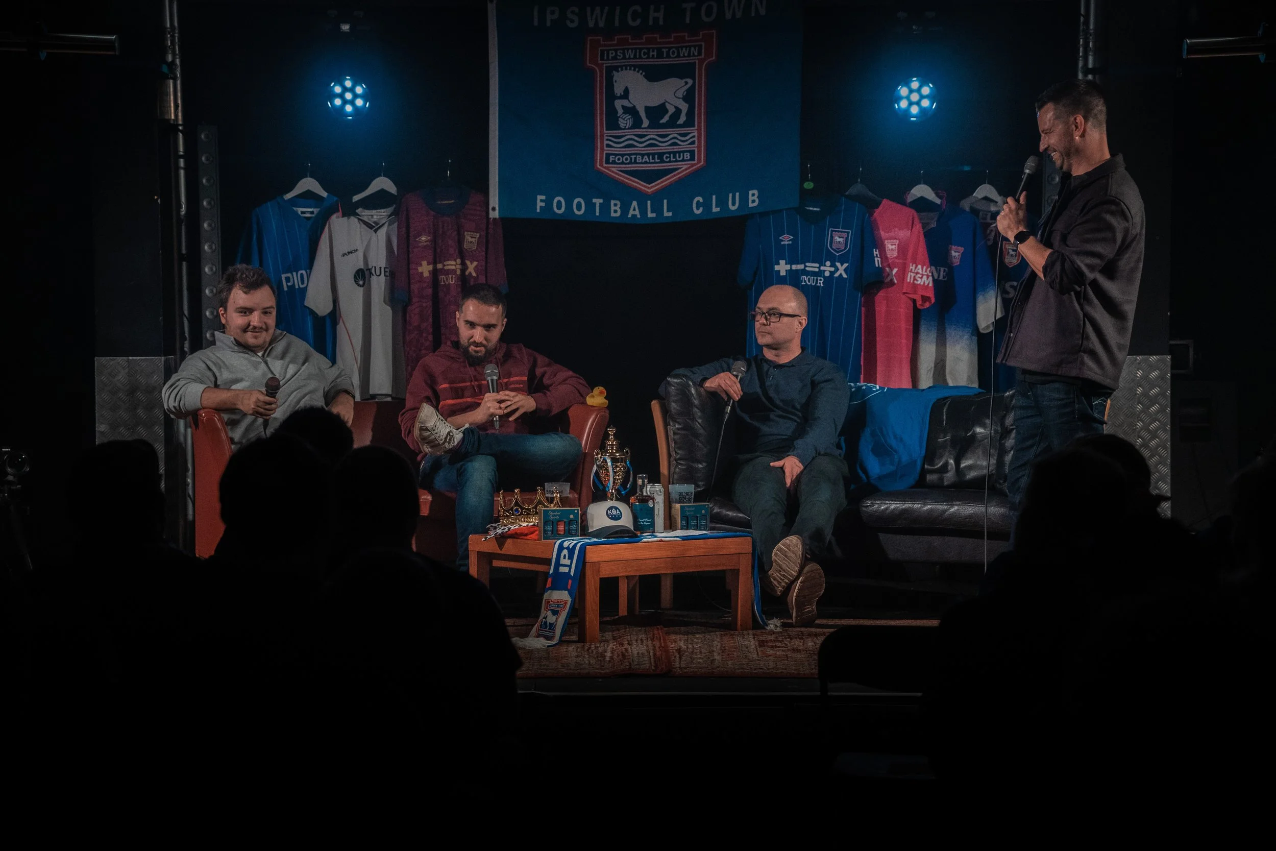 Four men sitting on chairs on a stage, engaging in a discussion or interview, with football jerseys and a banner for Ipswich Town Football Club hanging behind them. One man is standing and speaking into a microphone while the others listen.