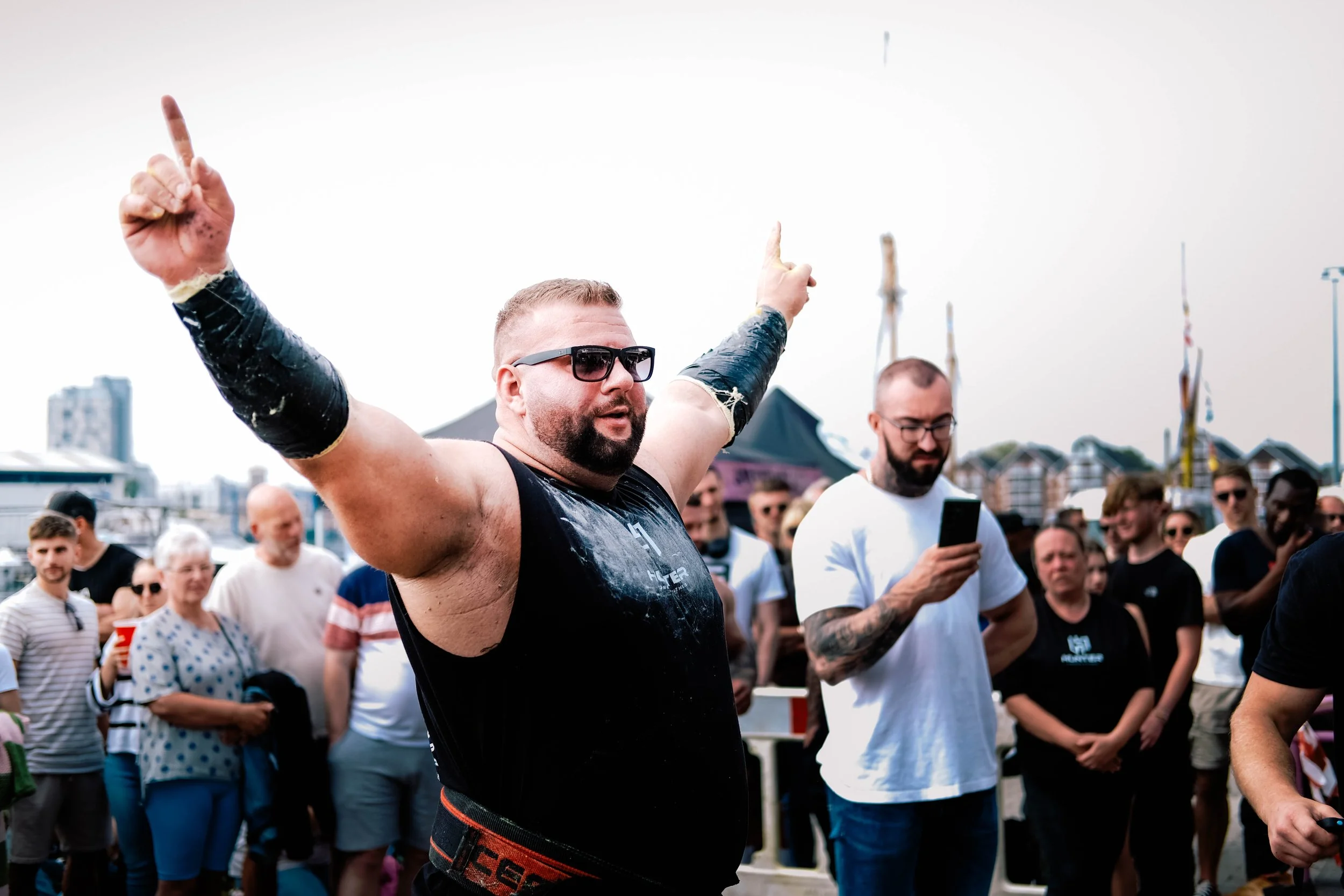 A man with sunglasses and a beard raising his arms, surrounded by a crowd outdoors on a cloudy day.