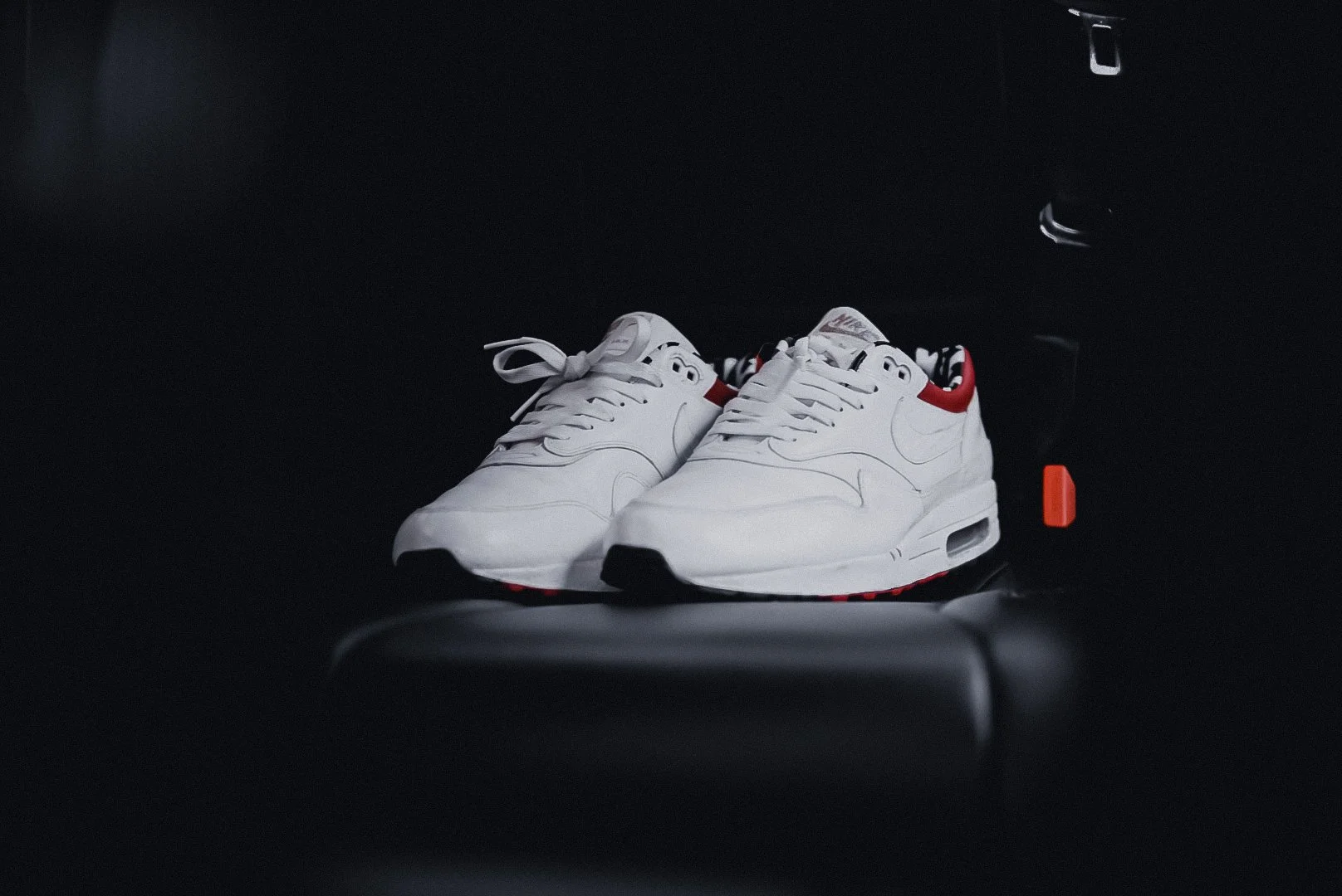 A pair of white Nike sneakers with red accents on a black surface, positioned against a black background with some orange and metal objects nearby.