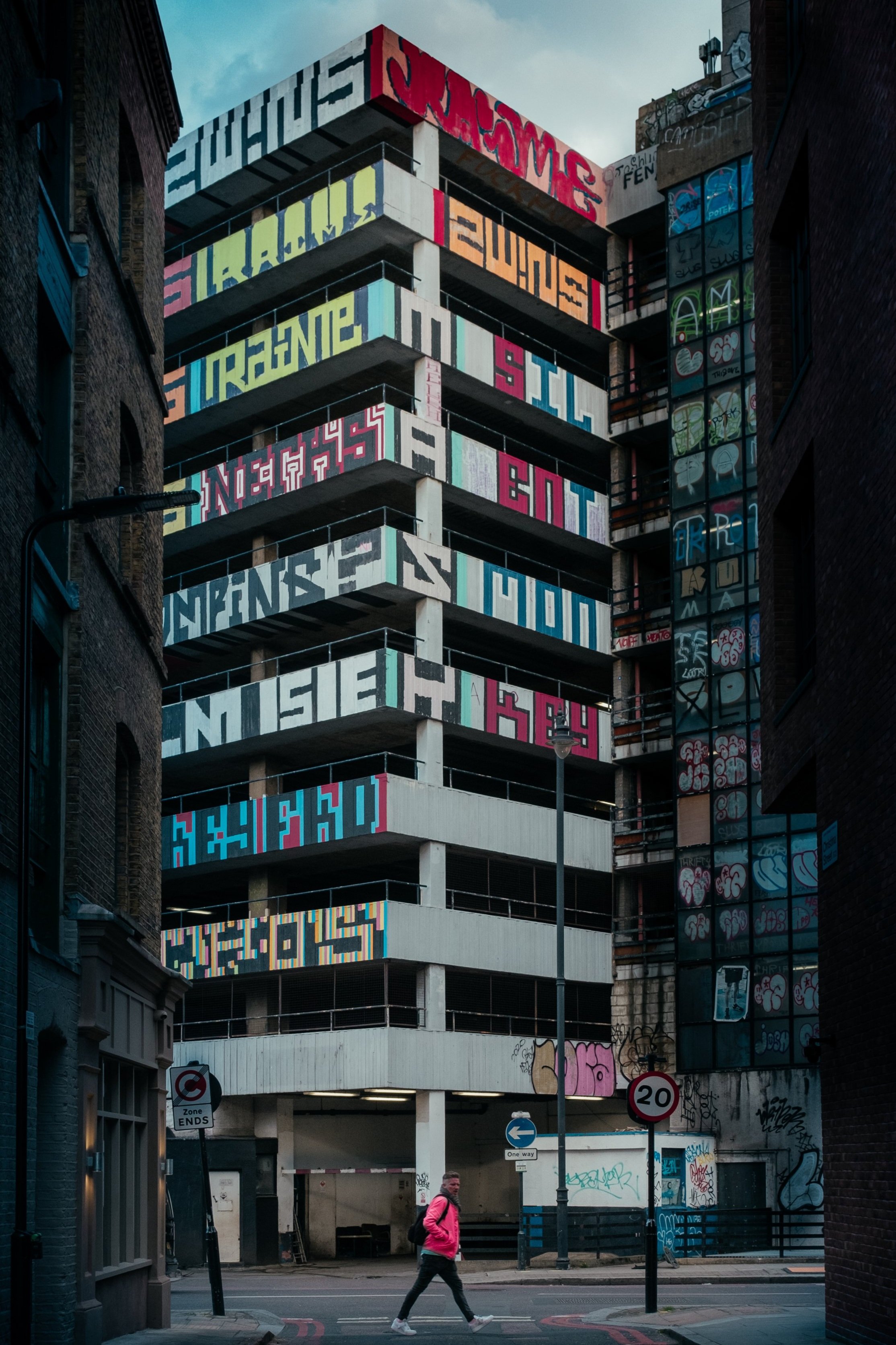 Multi-story parking garage with colorful graffiti-style lettering and artwork on the exterior walls, located in an urban area between brick buildings. A woman in a pink jacket is crossing the street in the foreground, and there are various street signs and a parking lot entrance below.
