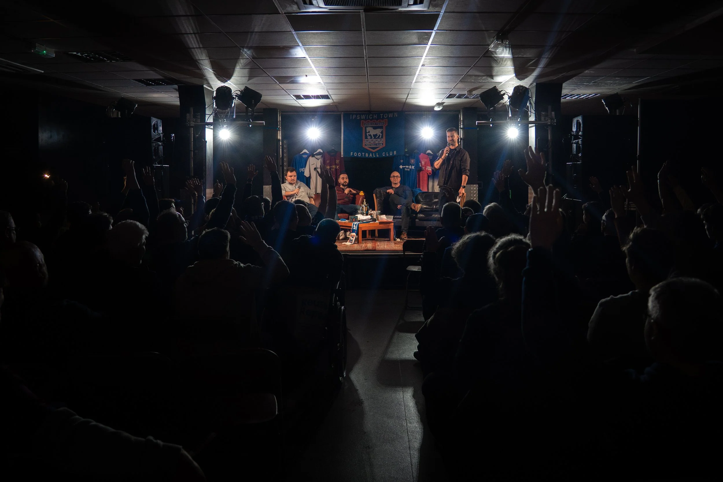 A panel discussion or talk taking place on a stage with four men seated and one man standing with a microphone. The audience has raised their hands. The background features sports jerseys and a banner for Ipswich Town Football Club. Stage lighting creates a silhouette effect of the viewers.