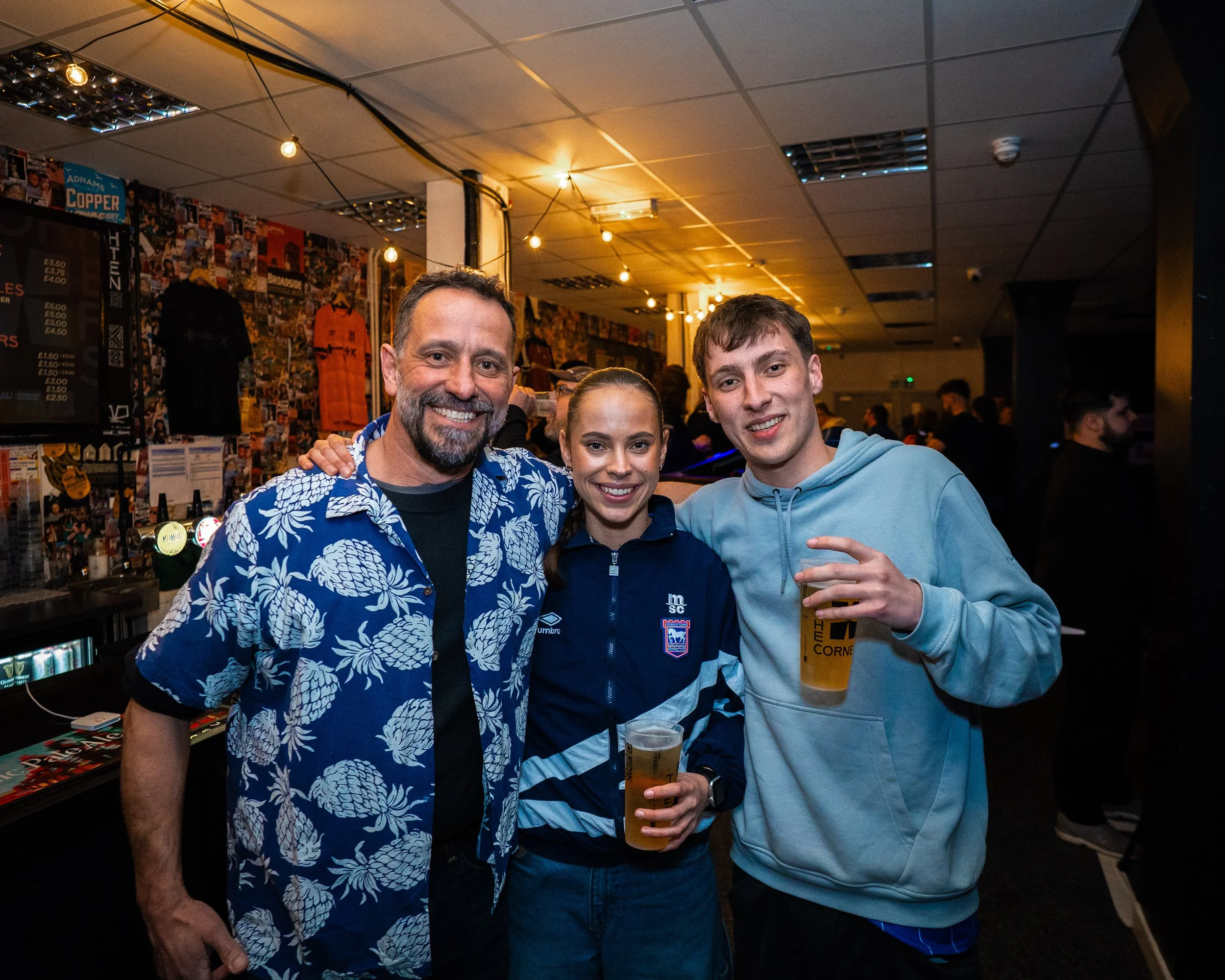 Three people smiling in a bar or pub, holding drinks, with a crowd in the background and decorated walls with posters and jerseys.